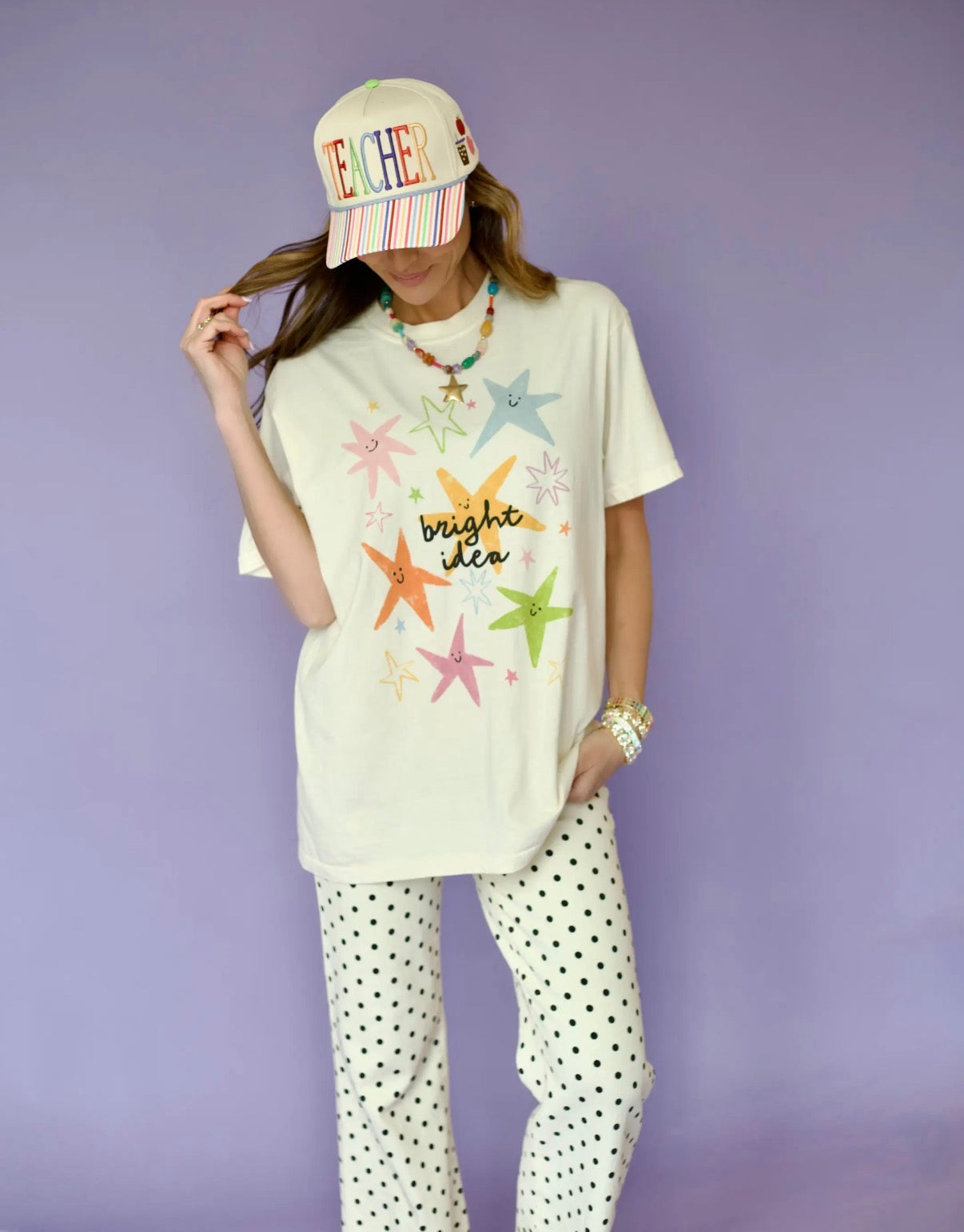 A woman modeling a cream-colored graphic t-shirt with colorful embroidered star designs and the text 'bright idea', paired with white pants featuring black polka dots, a striped baseball cap with 'FATHER' embroidered on it, and a colorful beaded necklace, set against a solid lavender background.