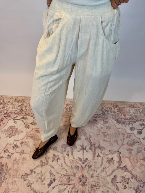A person wearing loose, light-colored linen pants with side pockets and dark flat shoes, standing on a patterned rug against a plain wall.