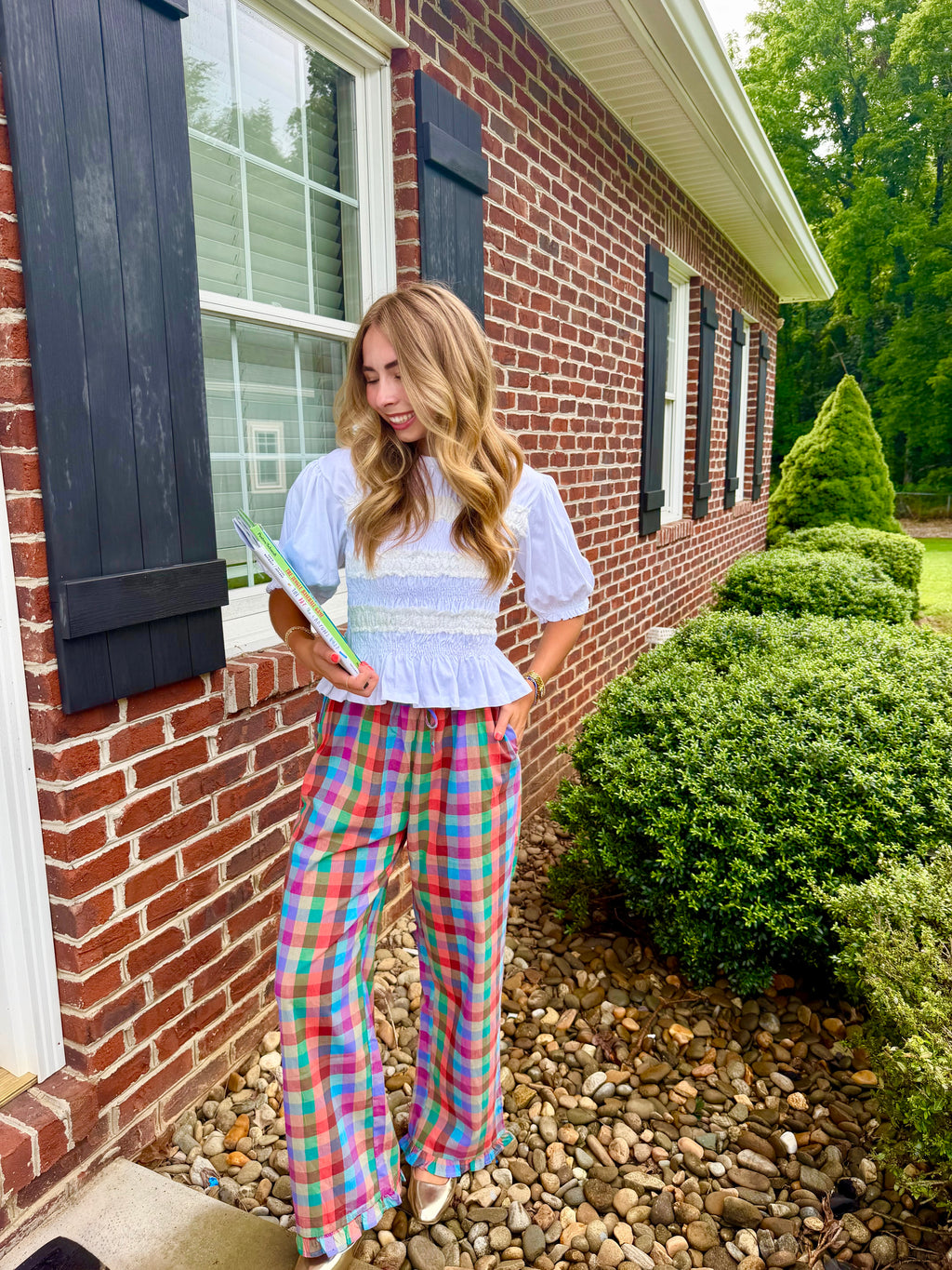 A woman stands outdoors in front of a red brick house, wearing a light blue and white ruffled blouse with colorful plaid wide-leg pants in pink, blue, green, and yellow. She is holding a green book or notebook.