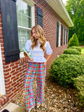 A woman stands outdoors in front of a red brick house, wearing a light blue and white ruffled blouse with colorful plaid wide-leg pants in pink, blue, green, and yellow. She is holding a green book or notebook.