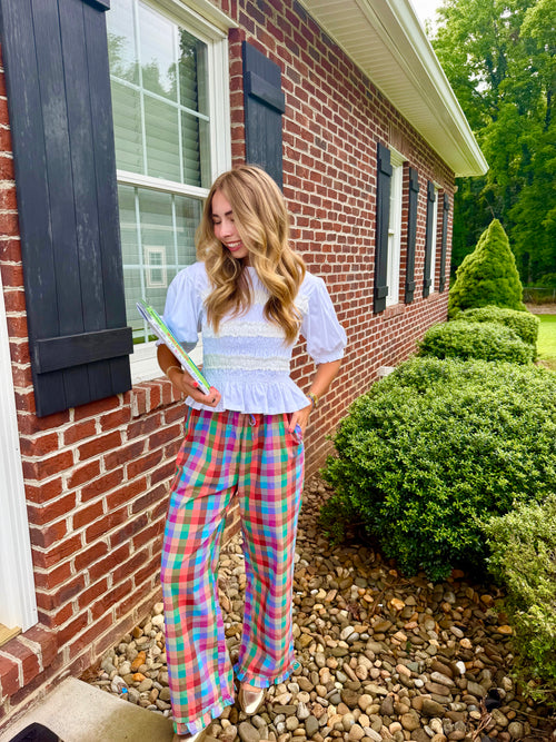 A woman stands outdoors in front of a red brick house, wearing a light blue and white ruffled blouse with colorful plaid wide-leg pants in pink, blue, green, and yellow. She is holding a green book or notebook.