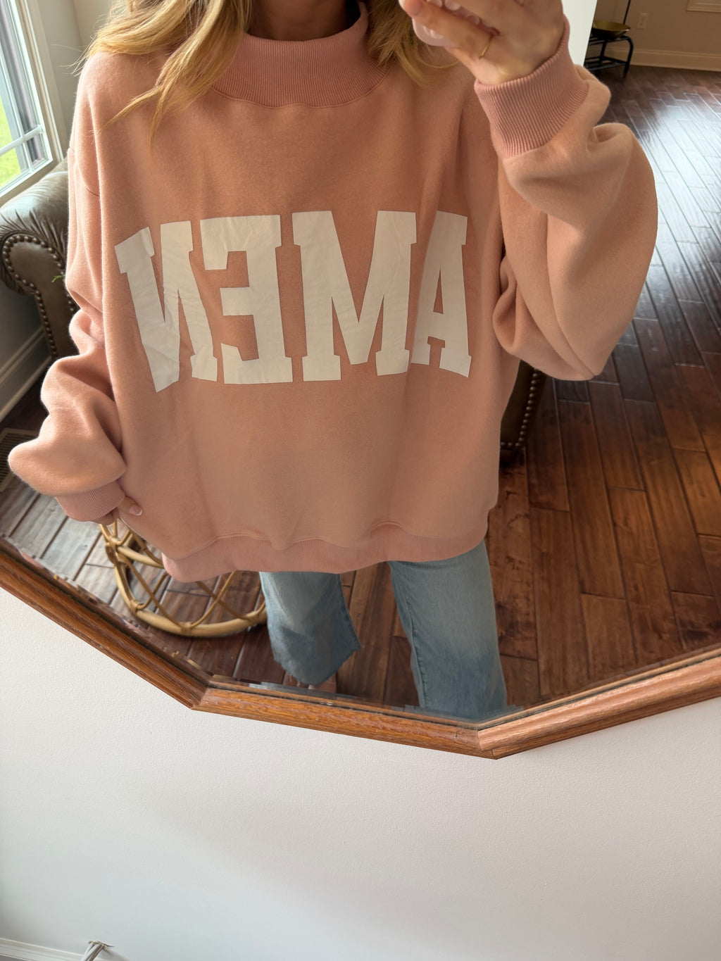 Person wearing a pink sweatshirt with 'amen' printed on it, standing in a room with wooden flooring.