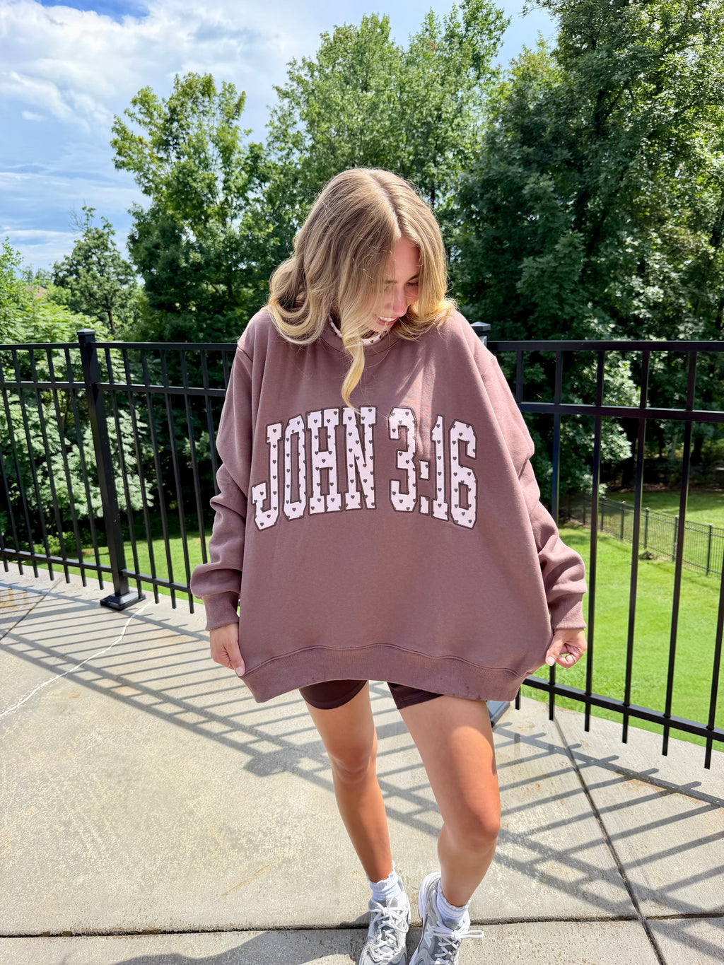 A woman wearing an oversized dusty rose sweatshirt with 'JOHN 3:16' printed in white block letters, standing on an outdoor patio with trees and a black metal fence in the background.