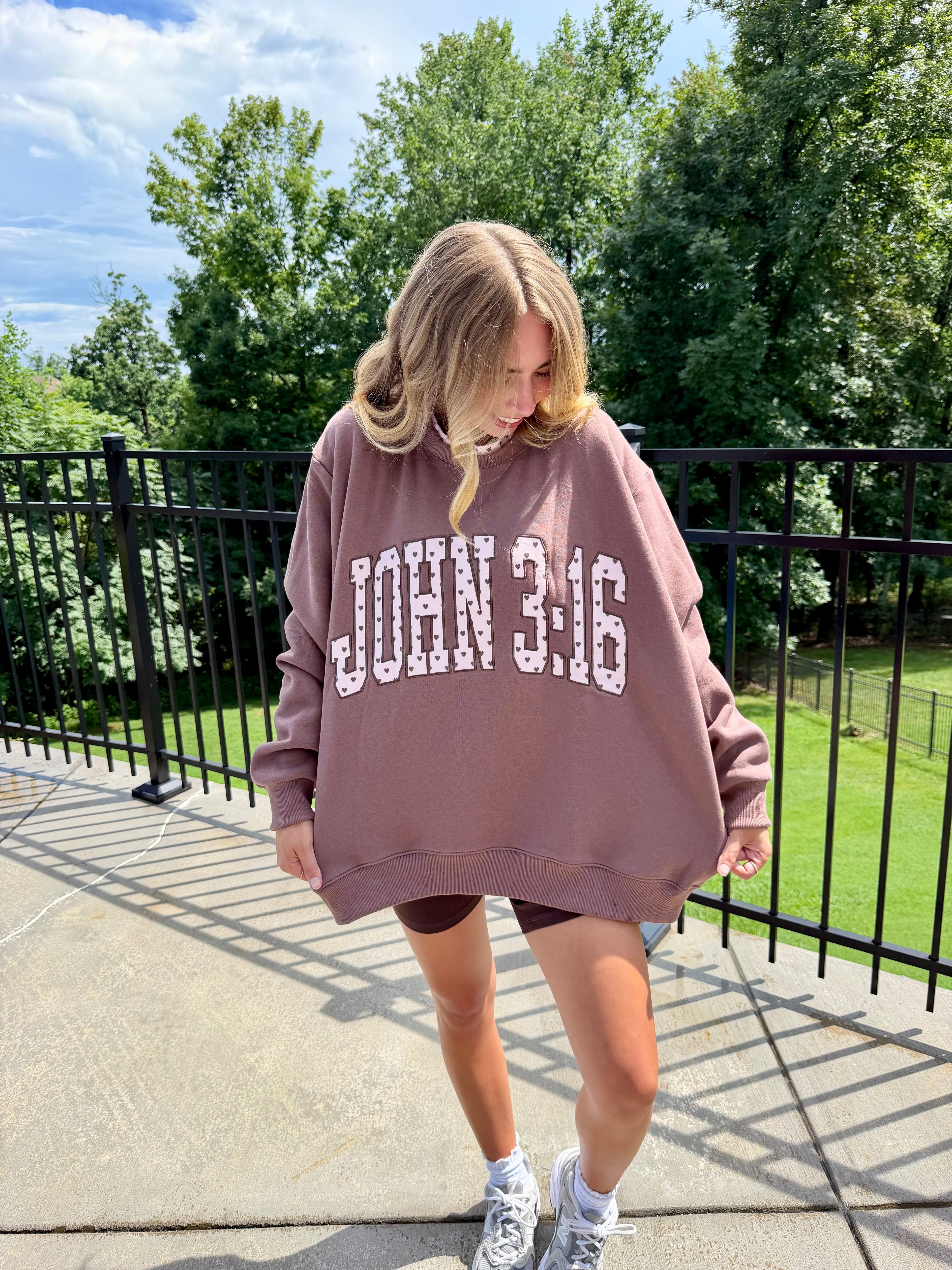 A woman wearing an oversized dusty rose sweatshirt with 'JOHN 3:16' printed in white block letters, standing on an outdoor patio with trees and a black metal fence in the background.