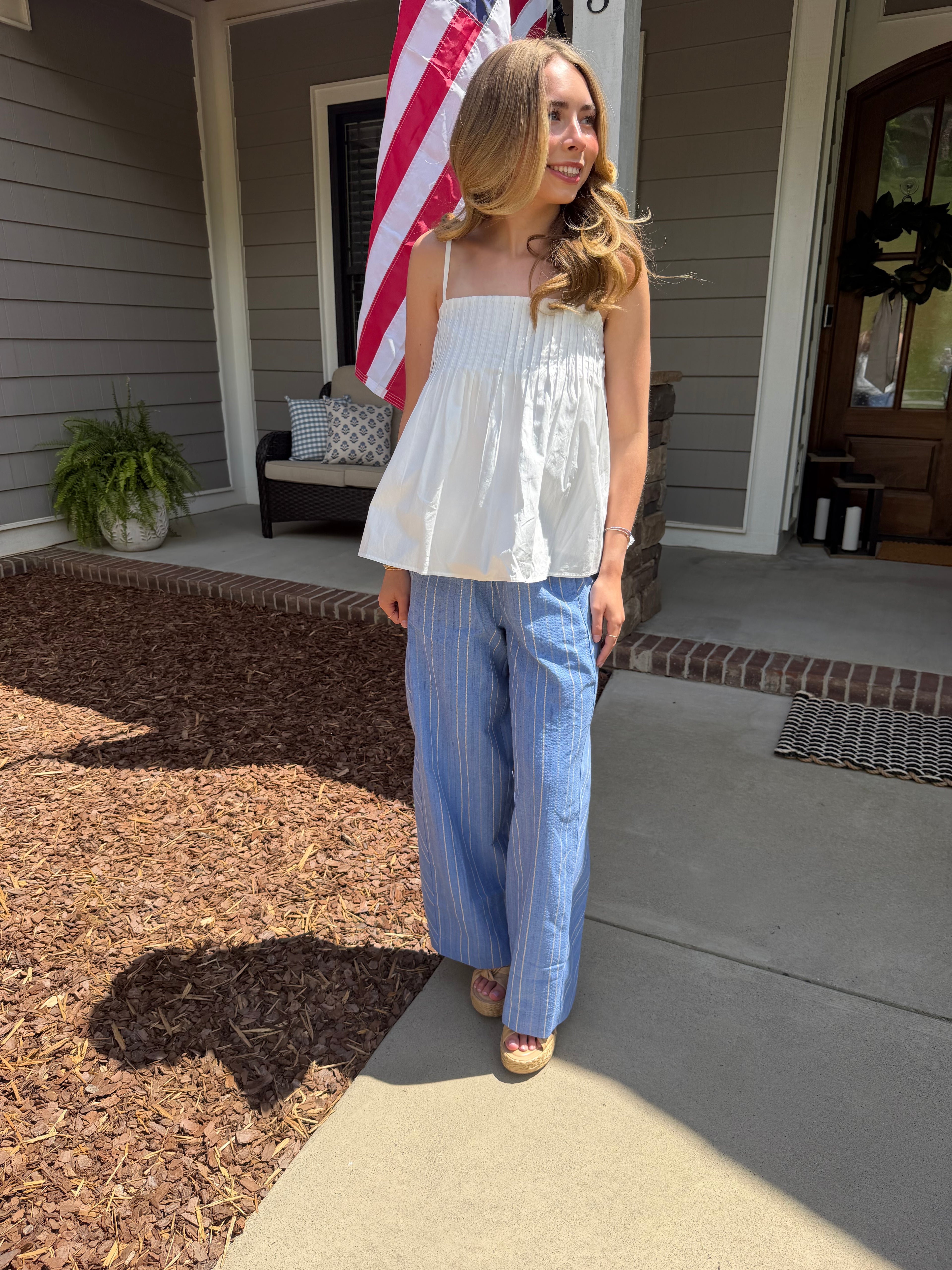 Woman standing on a porch wearing a white top and blue striped pants, with an American flag in the background.