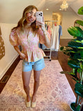 Woman taking a mirror selfie wearing a floral top and denim shorts in a home setting.
