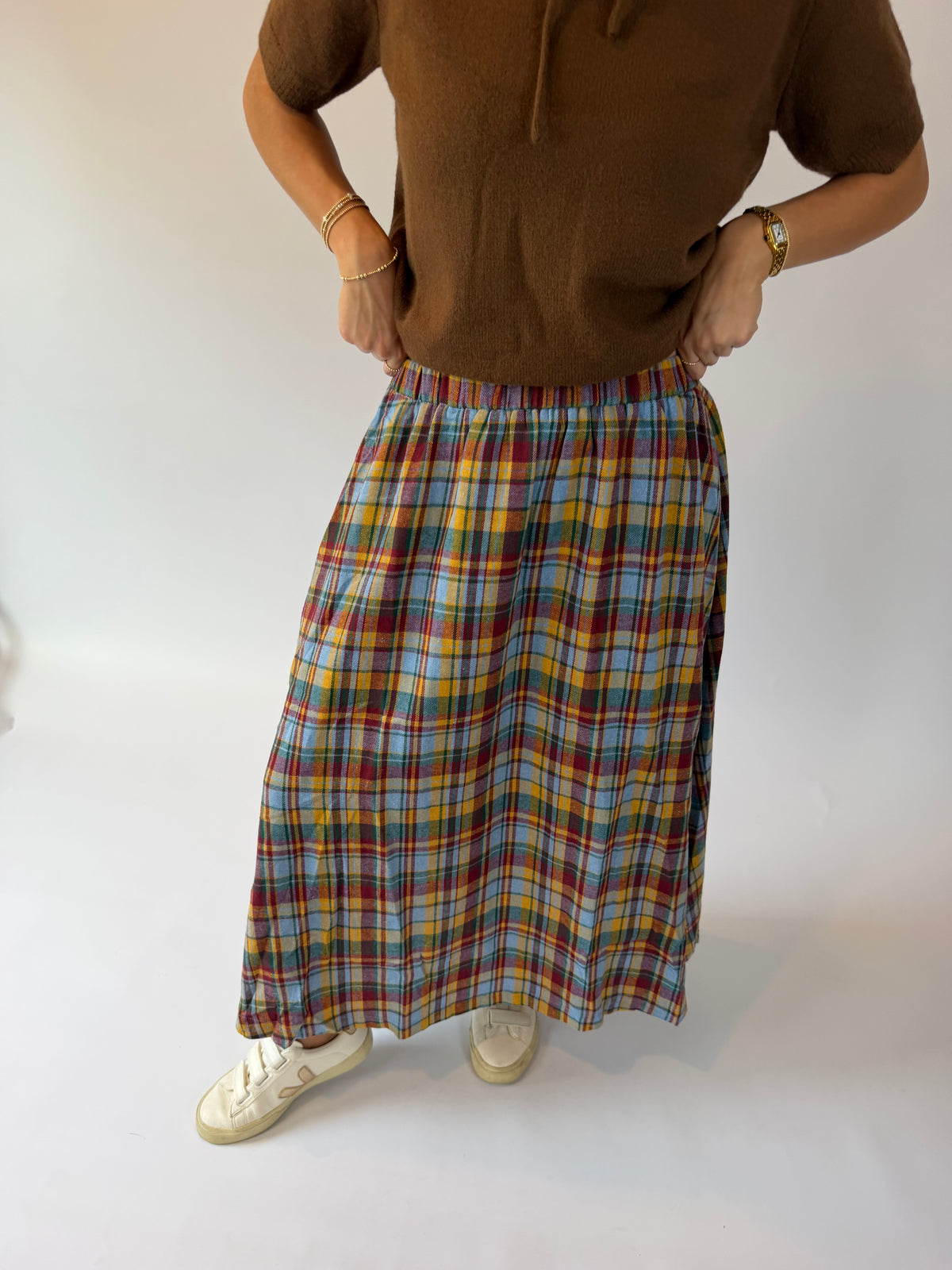 A person wearing a brown short-sleeve top and a long multicolor plaid skirt with white sneakers, standing against a plain white studio background.