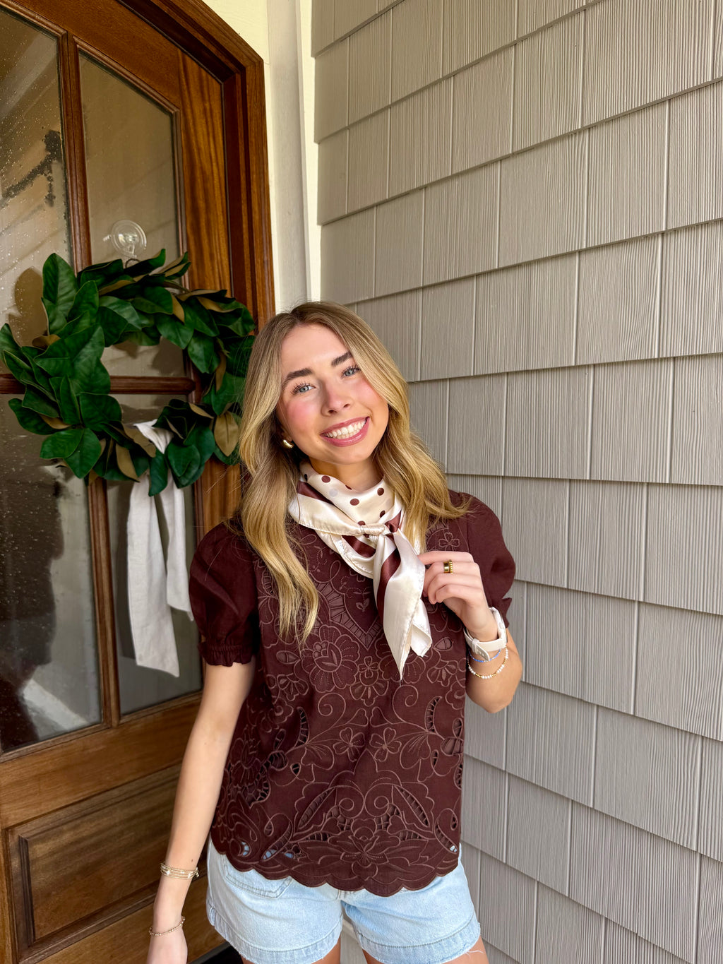 Woman standing outside a house with a wreath on the door, wearing a patterned top and scarf.
