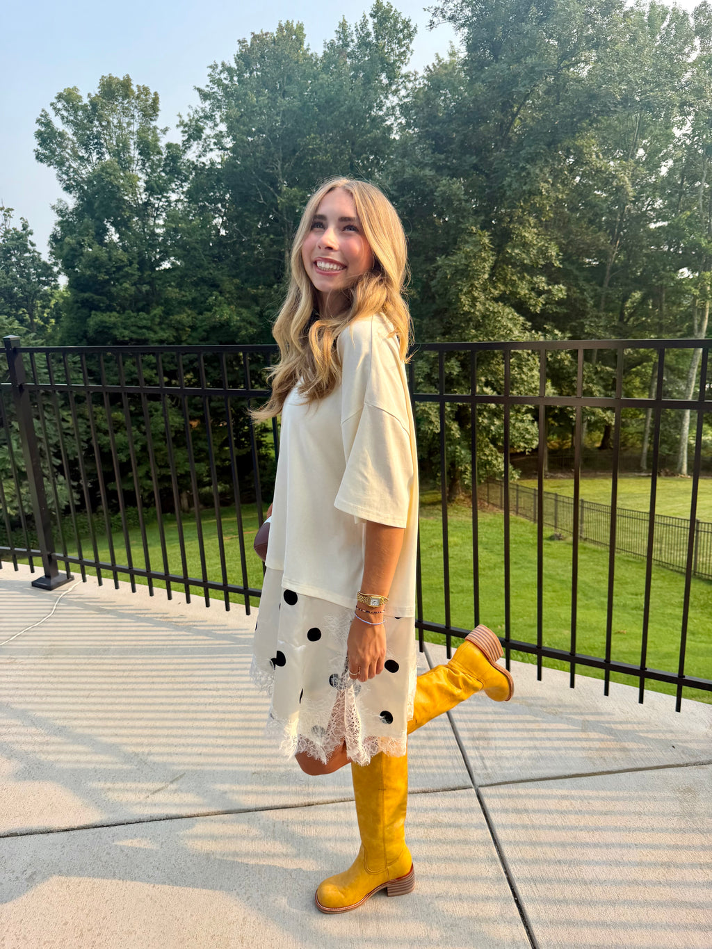 A woman stands on an outdoor concrete patio wearing a cream short-sleeve tunic dress with black polka dots and white lace trim, paired with bright yellow knee-high boots. She is posing with one leg raised in front of a black metal fence and green trees.