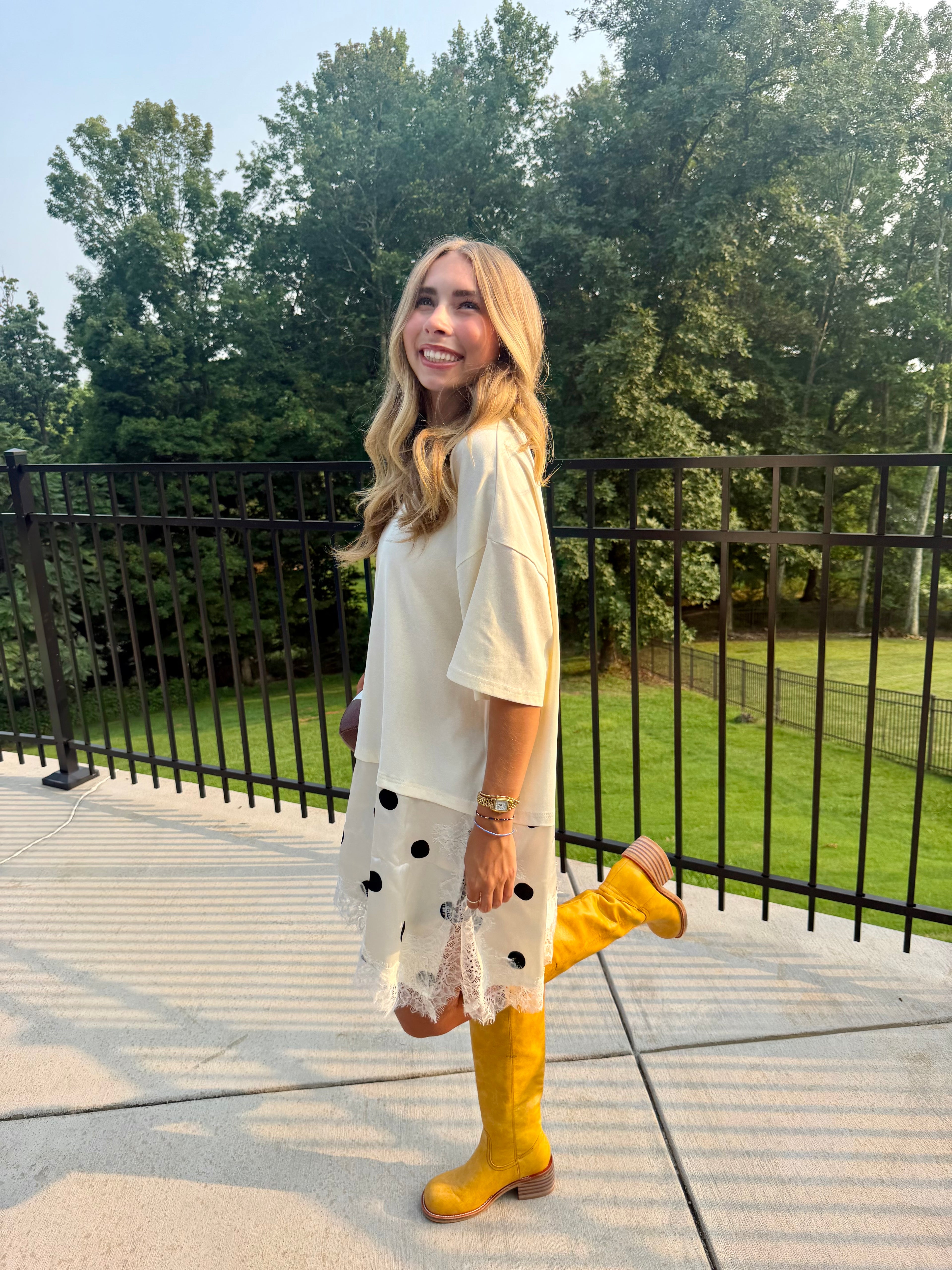 A woman stands on an outdoor concrete patio wearing a cream short-sleeve tunic dress with black polka dots and white lace trim, paired with bright yellow knee-high boots. She is posing with one leg raised in front of a black metal fence and green trees.