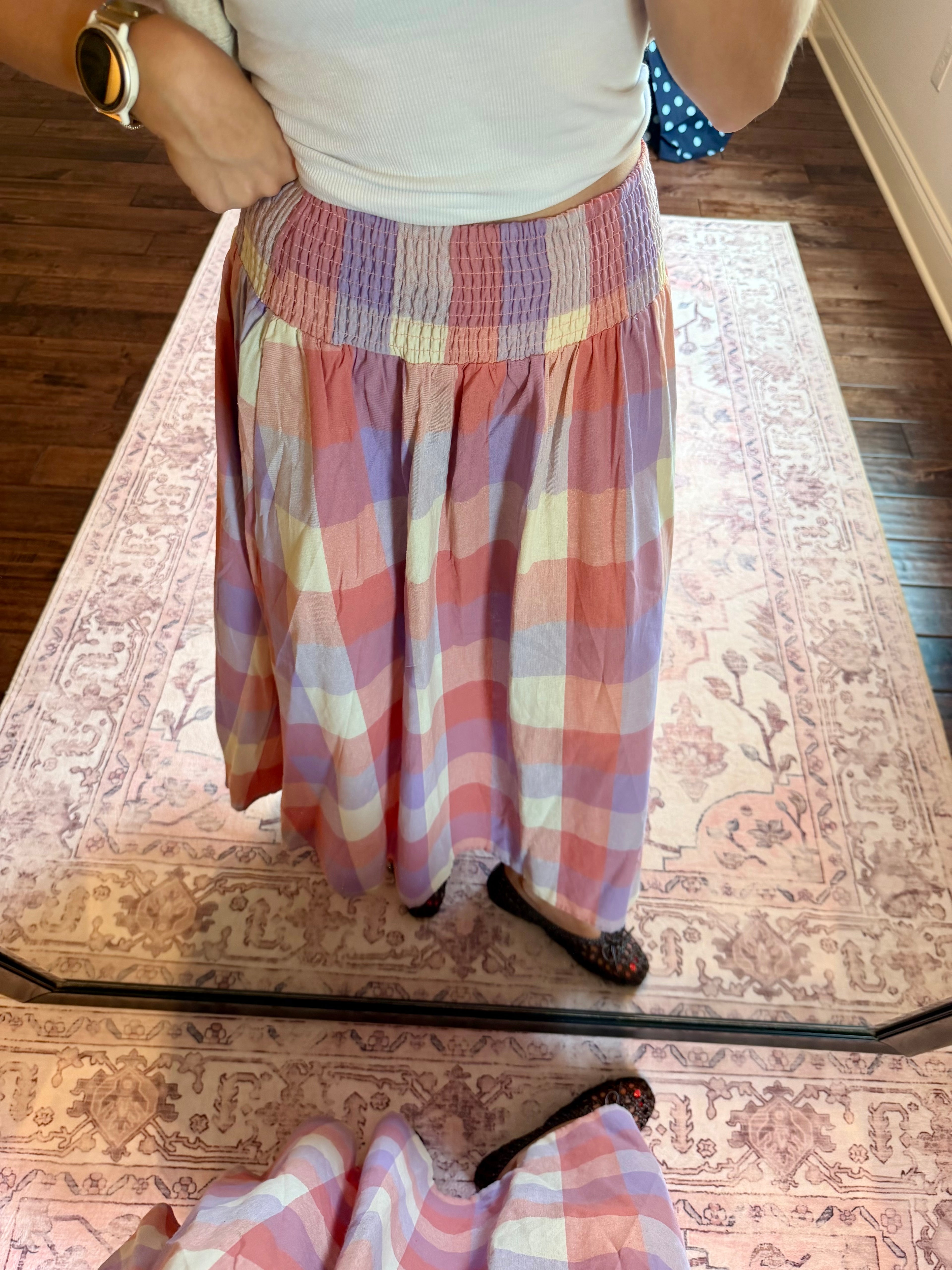 A person wearing a long, pleated maxi skirt with a pastel plaid pattern in pink, purple, yellow, and white, standing in front of a mirror on a patterned rug.