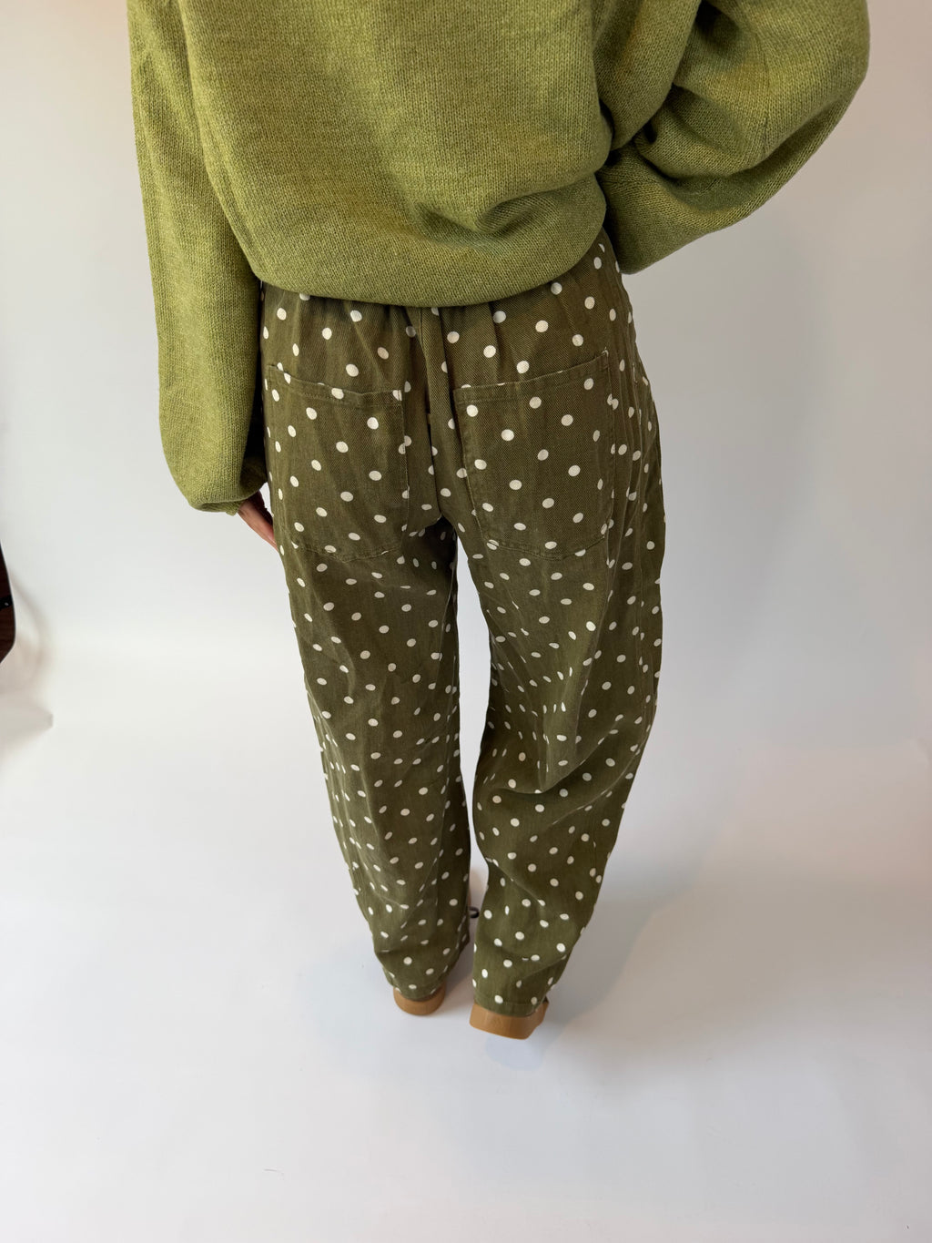 A person shown from the back wearing olive green pants with a white polka-dot pattern and a matching olive green sweater, standing against a plain white studio background.