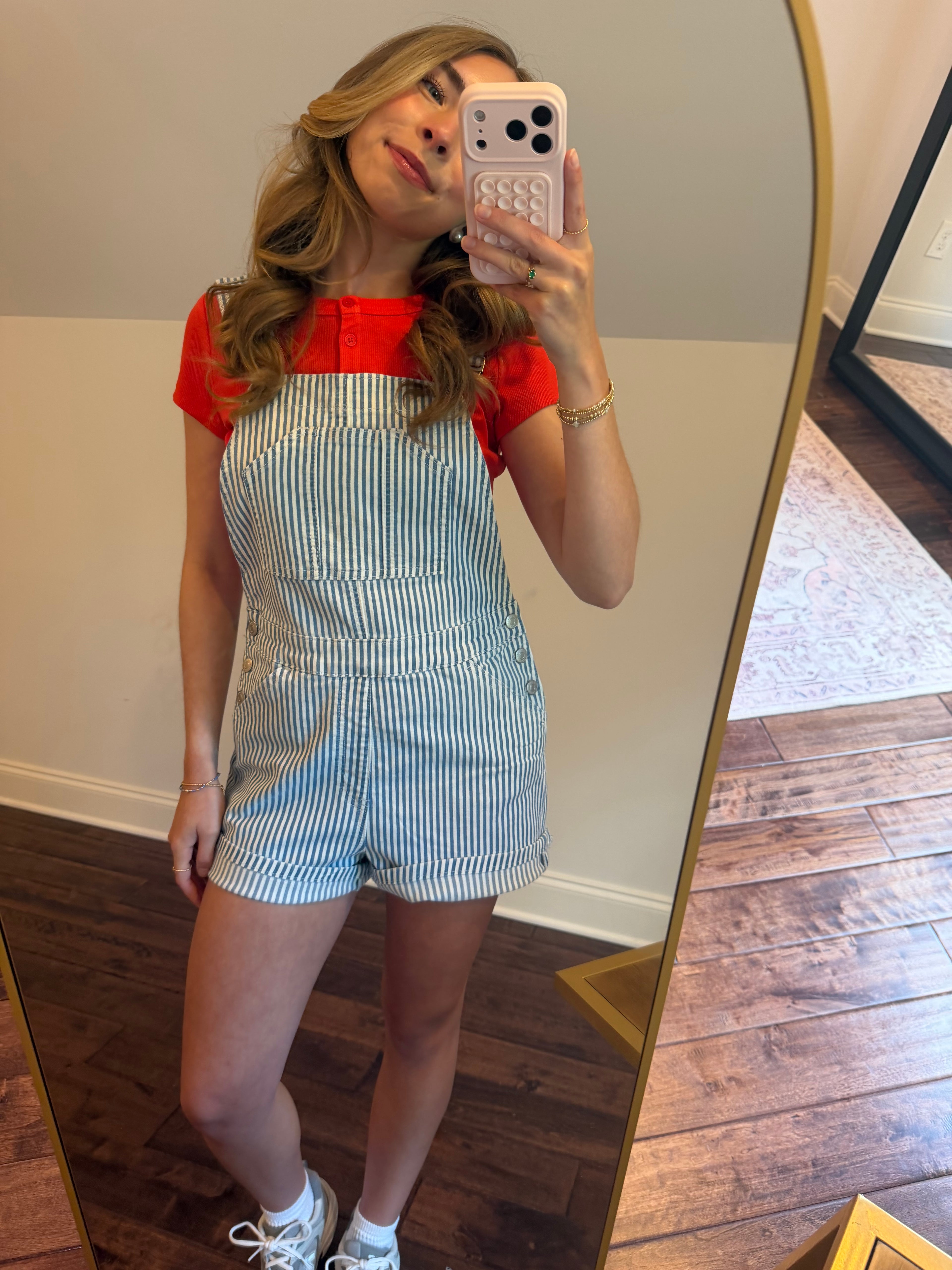 Woman taking a mirror selfie wearing a red top and blue striped overalls.