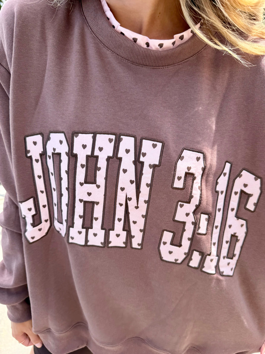 A close-up of a person wearing a dusty mauve crewneck sweatshirt with 'JOHN 3:16' printed in white varsity letters with small heart patterns, layered over a pink and white patterned collar.