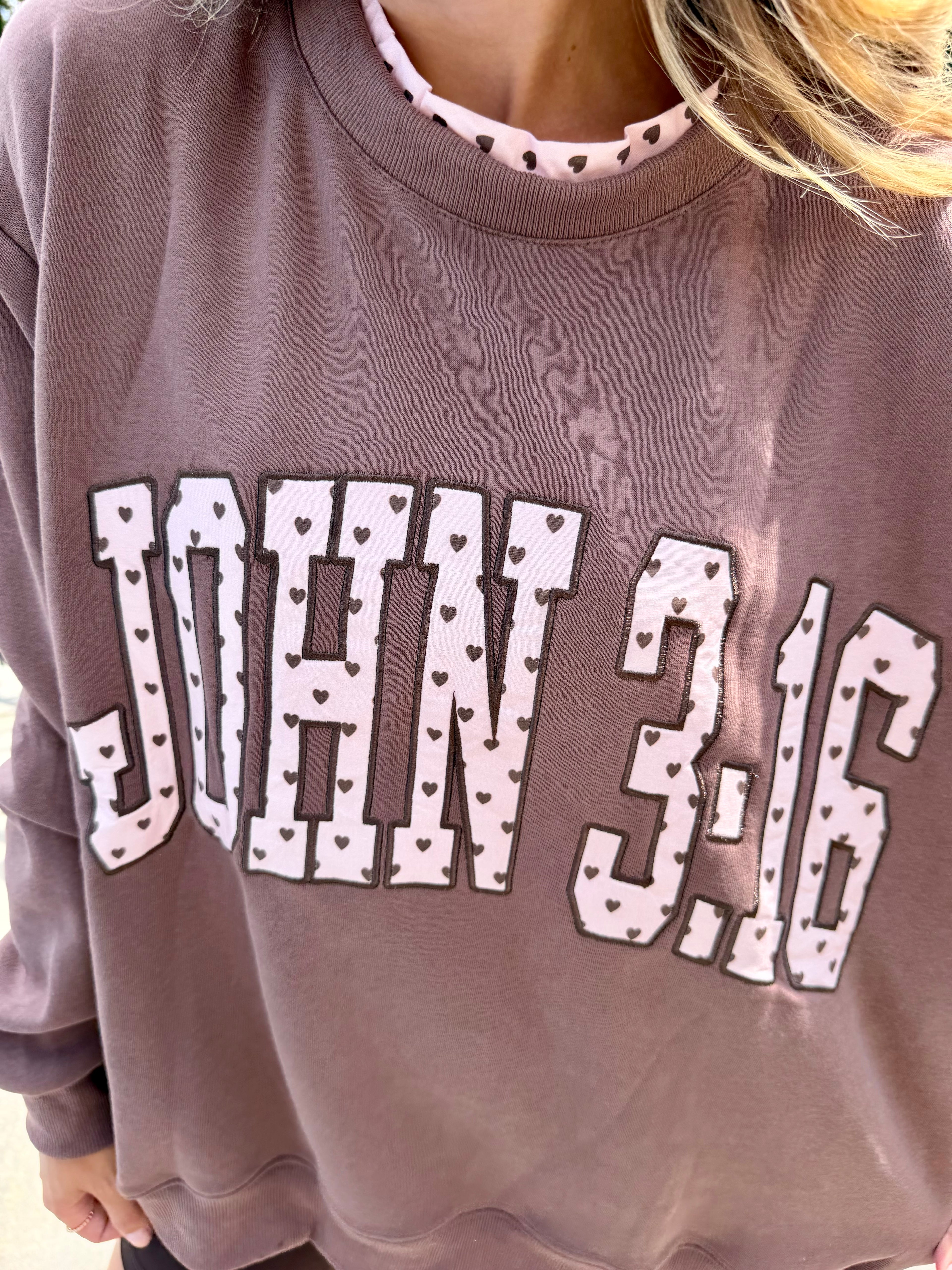 A close-up of a person wearing a dusty mauve crewneck sweatshirt with 'JOHN 3:16' printed in white varsity letters with small heart patterns, layered over a pink and white patterned collar.