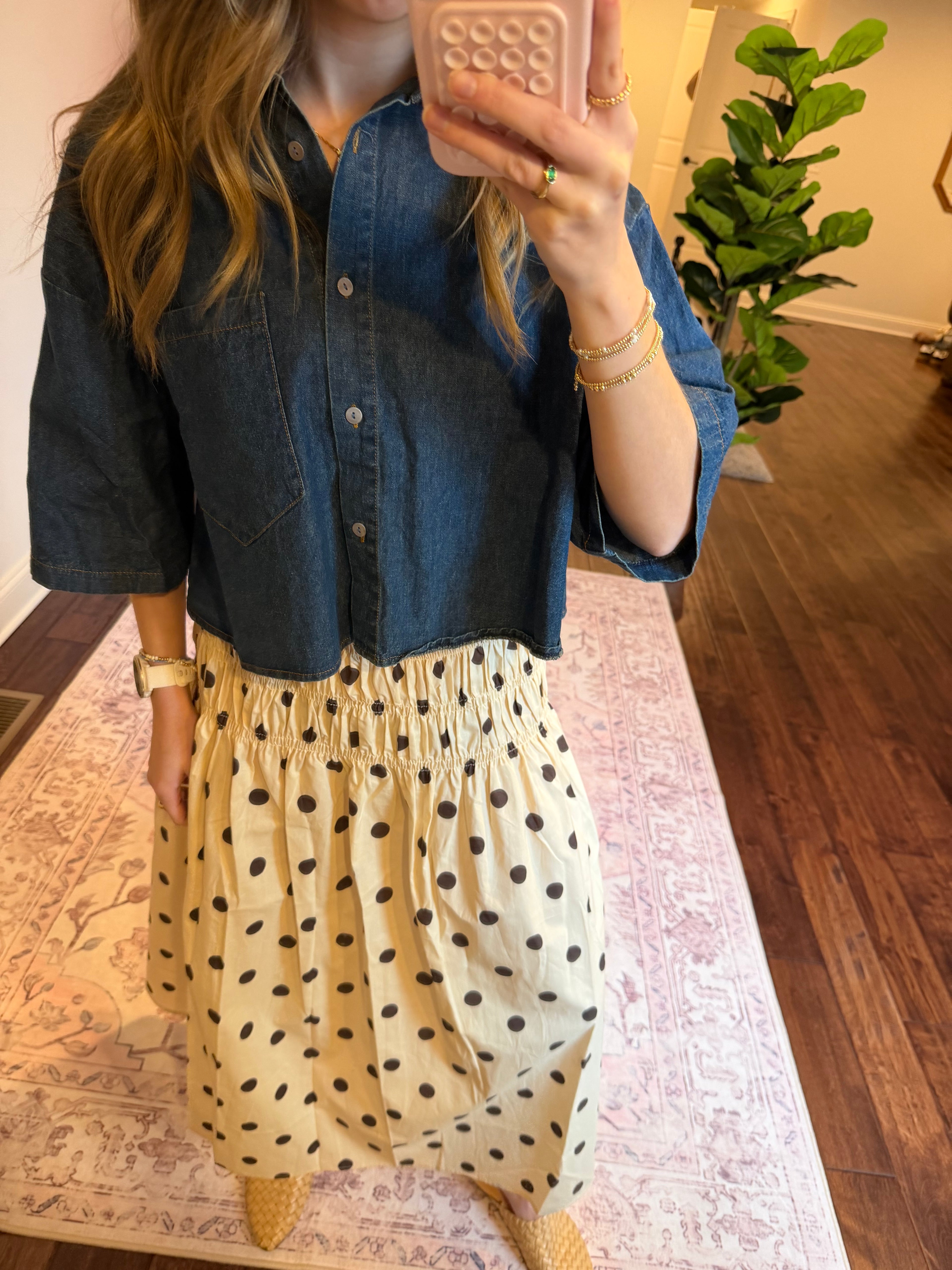 Woman taking a mirror selfie wearing a denim shirt and polka dot skirt in a room with a plant and bed.