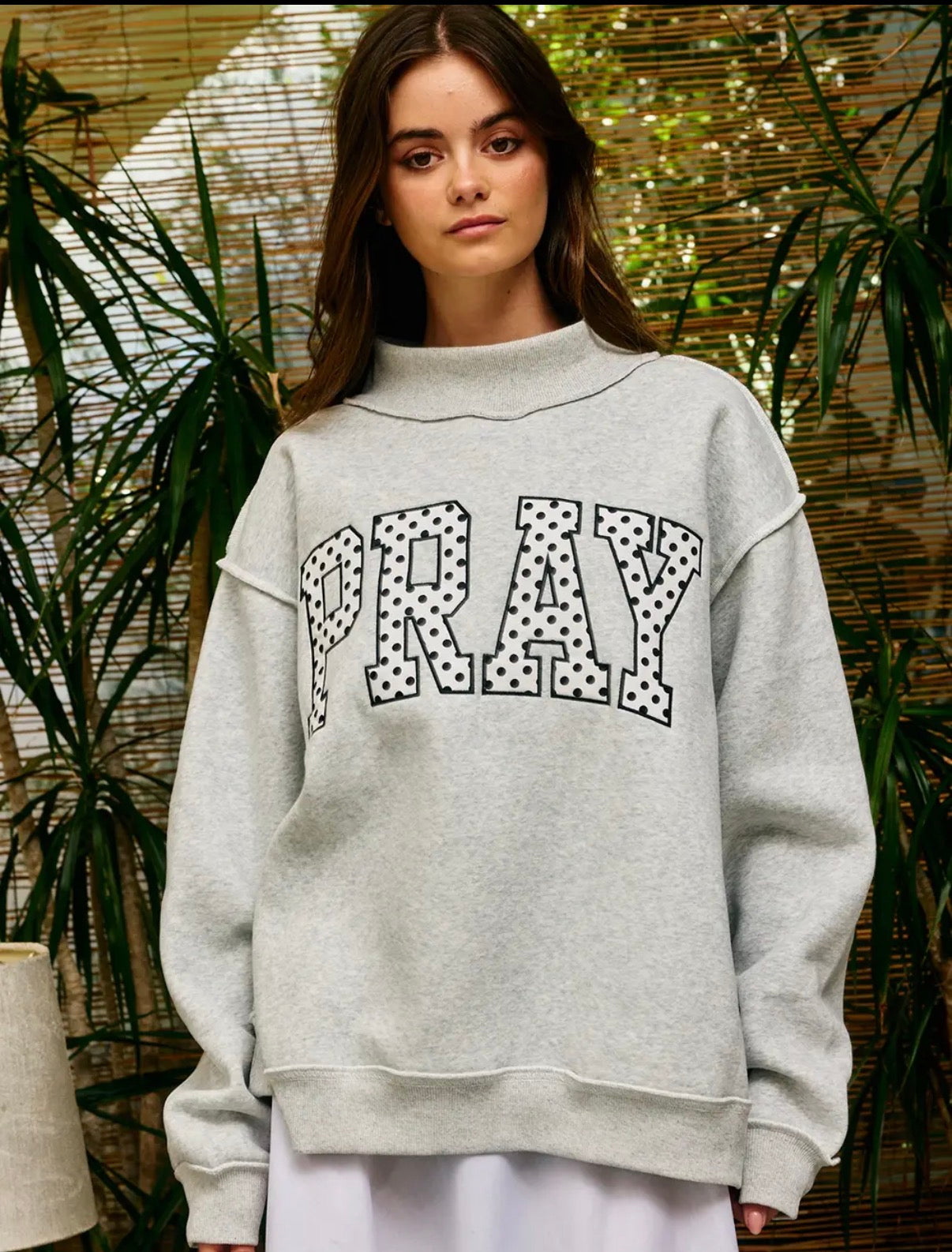 A woman wearing a light gray heather sweatshirt with the word 'PRAY' in large black-and-white polka dot outlined letters across the chest, posed in front of a bamboo screen and plants.