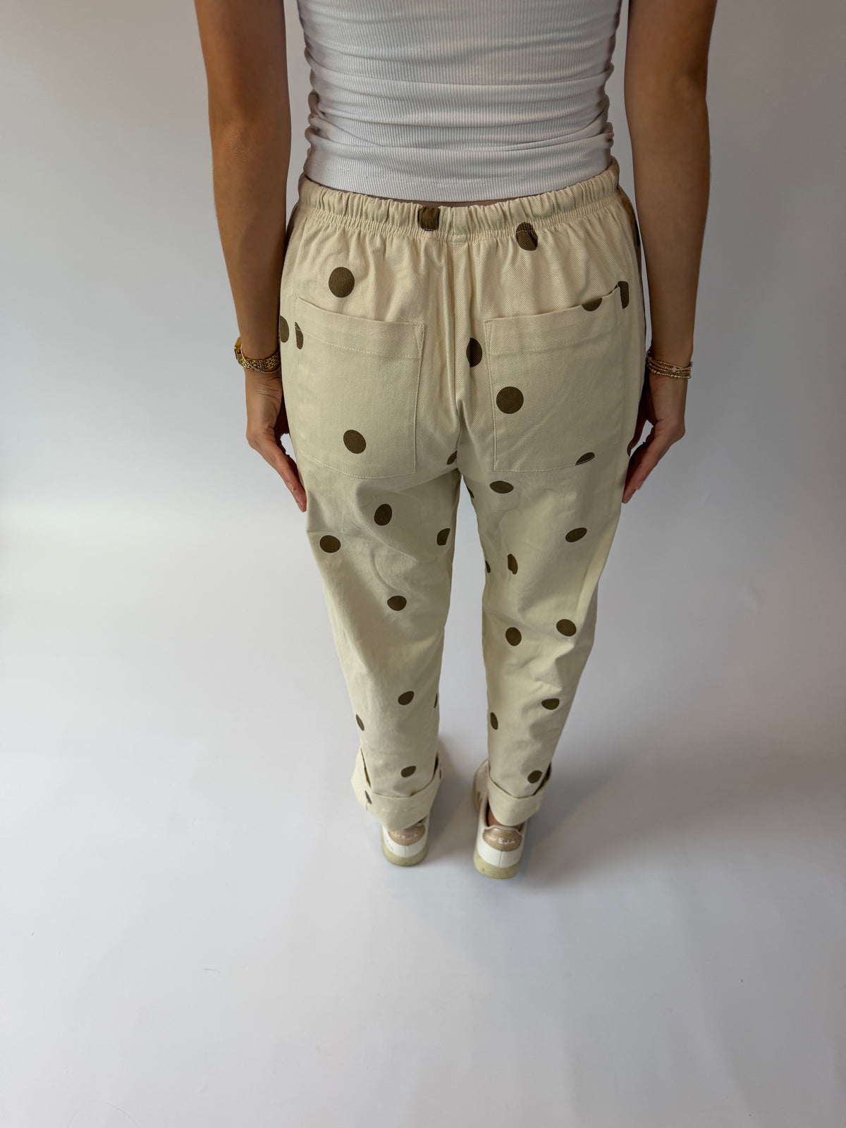 A person shown from the back wearing cream-colored pants with dark brown polka dots, paired with a white ribbed top and white sneakers, standing against a plain light gray studio background.