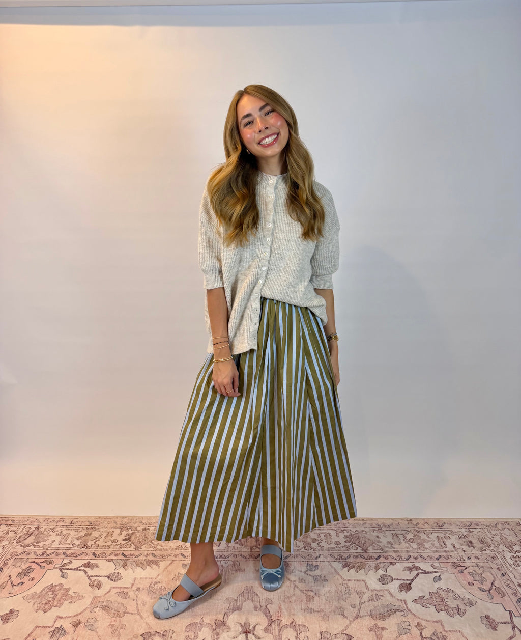 A woman stands indoors wearing a light gray knit top and a long skirt with vertical olive green and white stripes. She is posed on a patterned pink rug against a plain light gray wall.