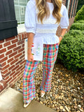 A woman wearing a white smocked blouse and colorful plaid wide-leg pants, standing outdoors next to a brick wall and gravel path.
