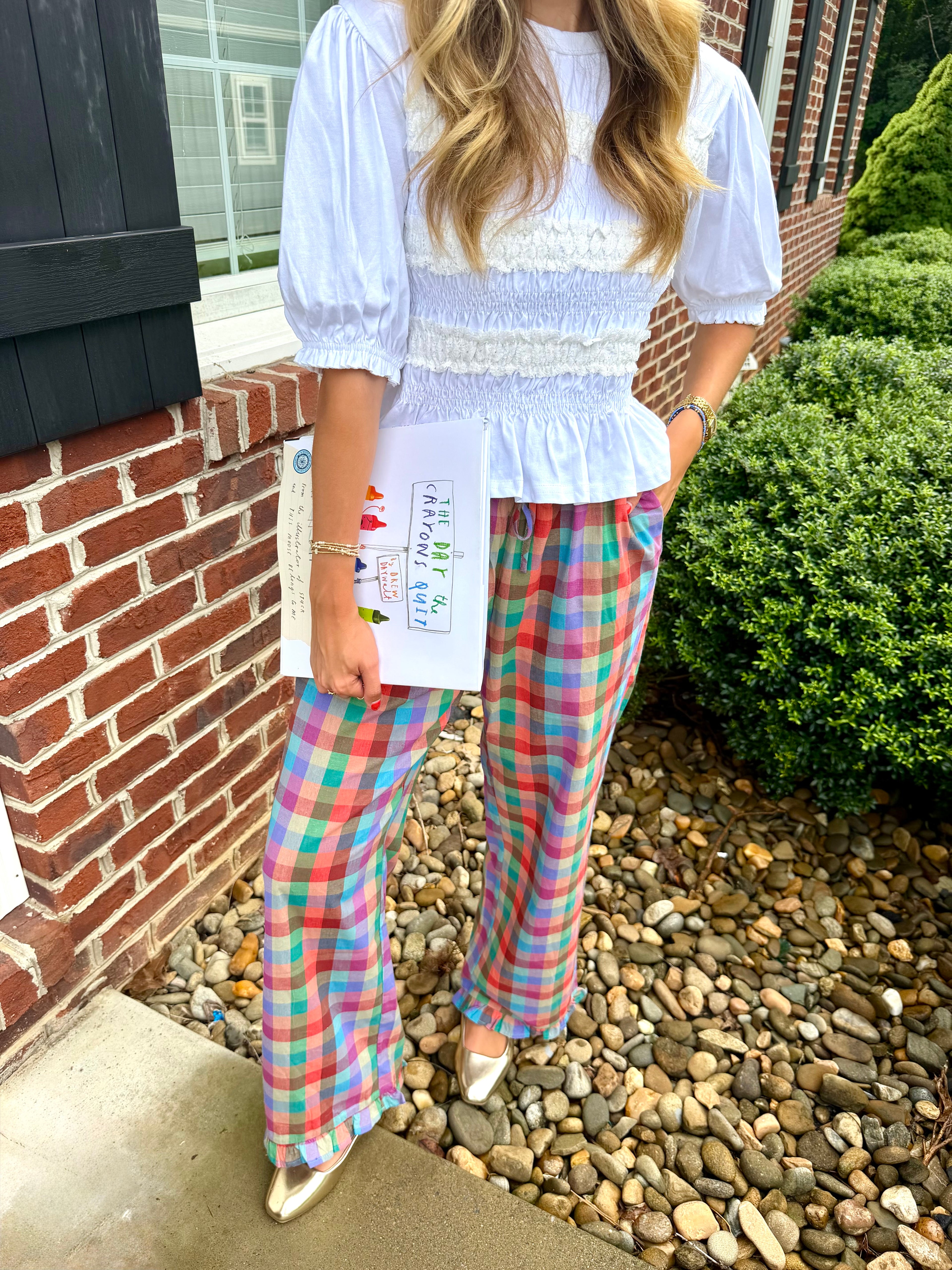 A woman wearing a white smocked blouse and colorful plaid wide-leg pants, standing outdoors next to a brick wall and gravel path.