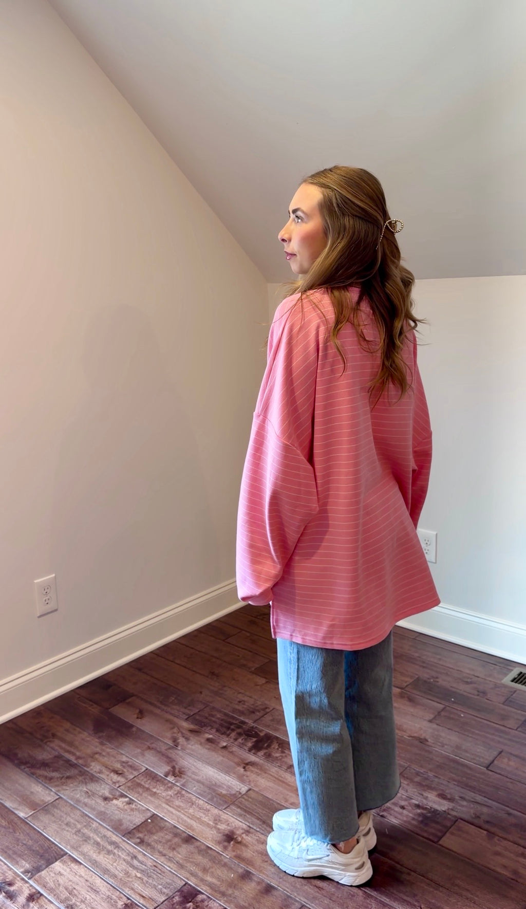 Person wearing a pink shirt and blue jeans standing in a room with wooden flooring and white walls.