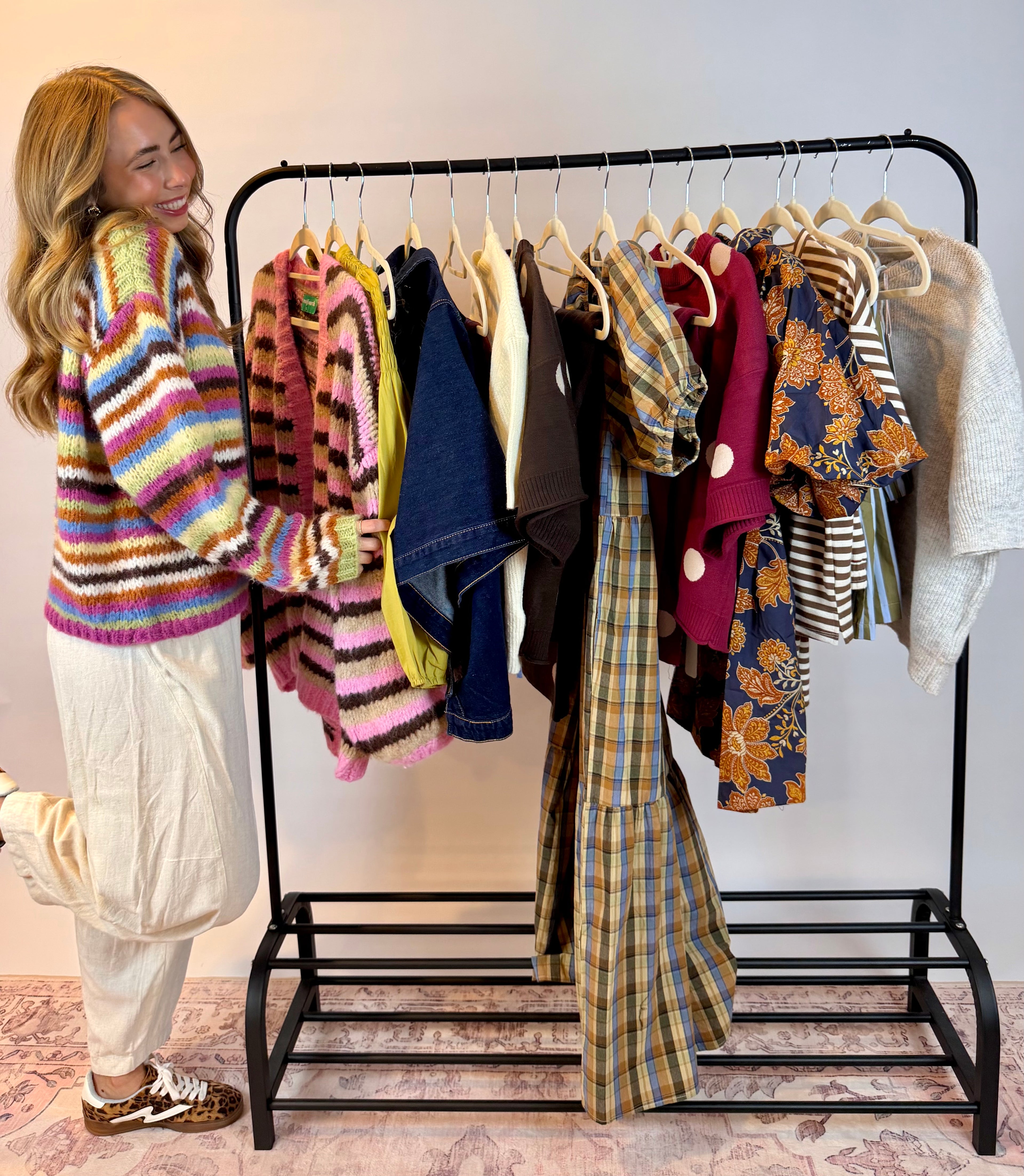 A woman stands beside a black clothing rack filled with multiple colorful knit and patterned tops, in a home setting with a patterned rug and light wall.