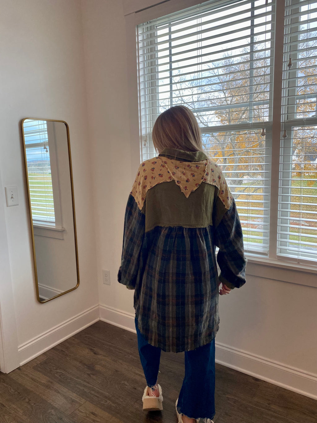 Person wearing a patterned jacket standing in a room with a window and mirror.