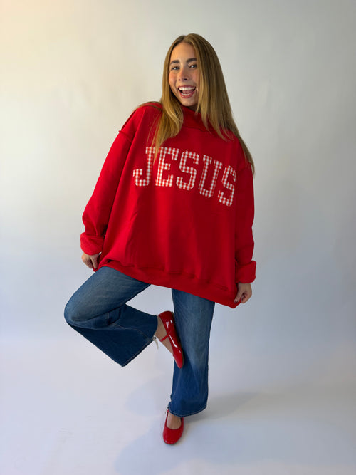A woman posing in a red oversized sweatshirt with 'JESUS' written across the chest in white rhinestone letters, paired with blue jeans and red shoes, against a plain white studio background.