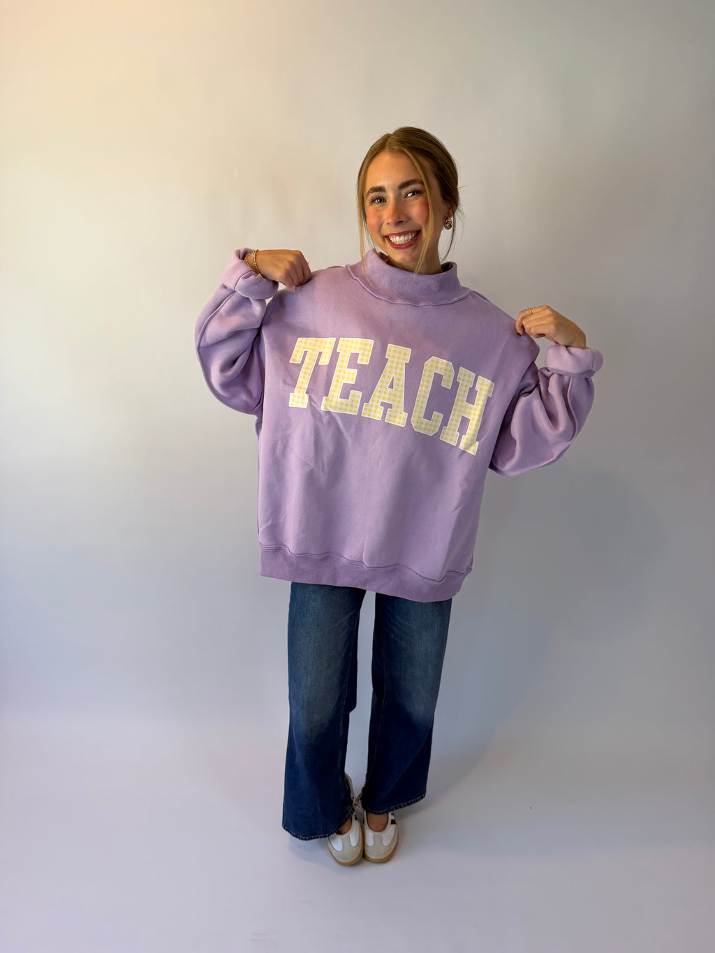 A woman wearing a lavender purple sweatshirt with 'TEACH' printed in gold block letters, paired with dark blue jeans and white sneakers, posing against a plain light gray background.