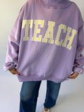 A person wearing an oversized lavender sweatshirt with the word 'TEACH' printed across the back in large yellow-and-white checkered letters, paired with blue jeans.