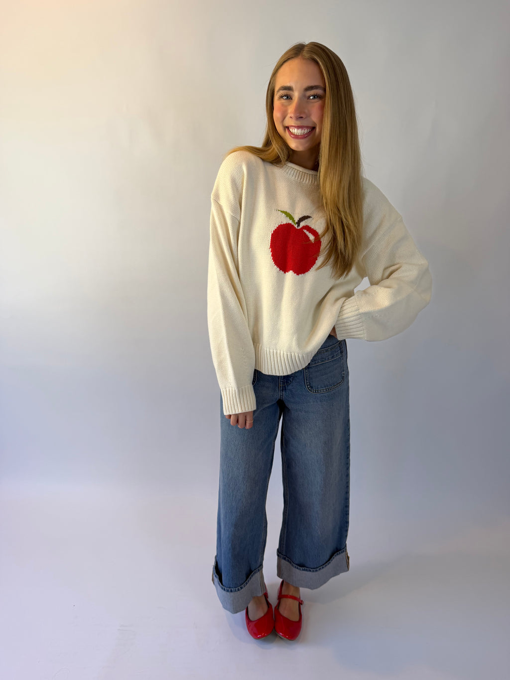 A woman modeling a cream-colored knit sweater featuring a red apple graphic on the front, paired with wide-leg blue jeans and red shoes, standing against a plain light gray studio background.