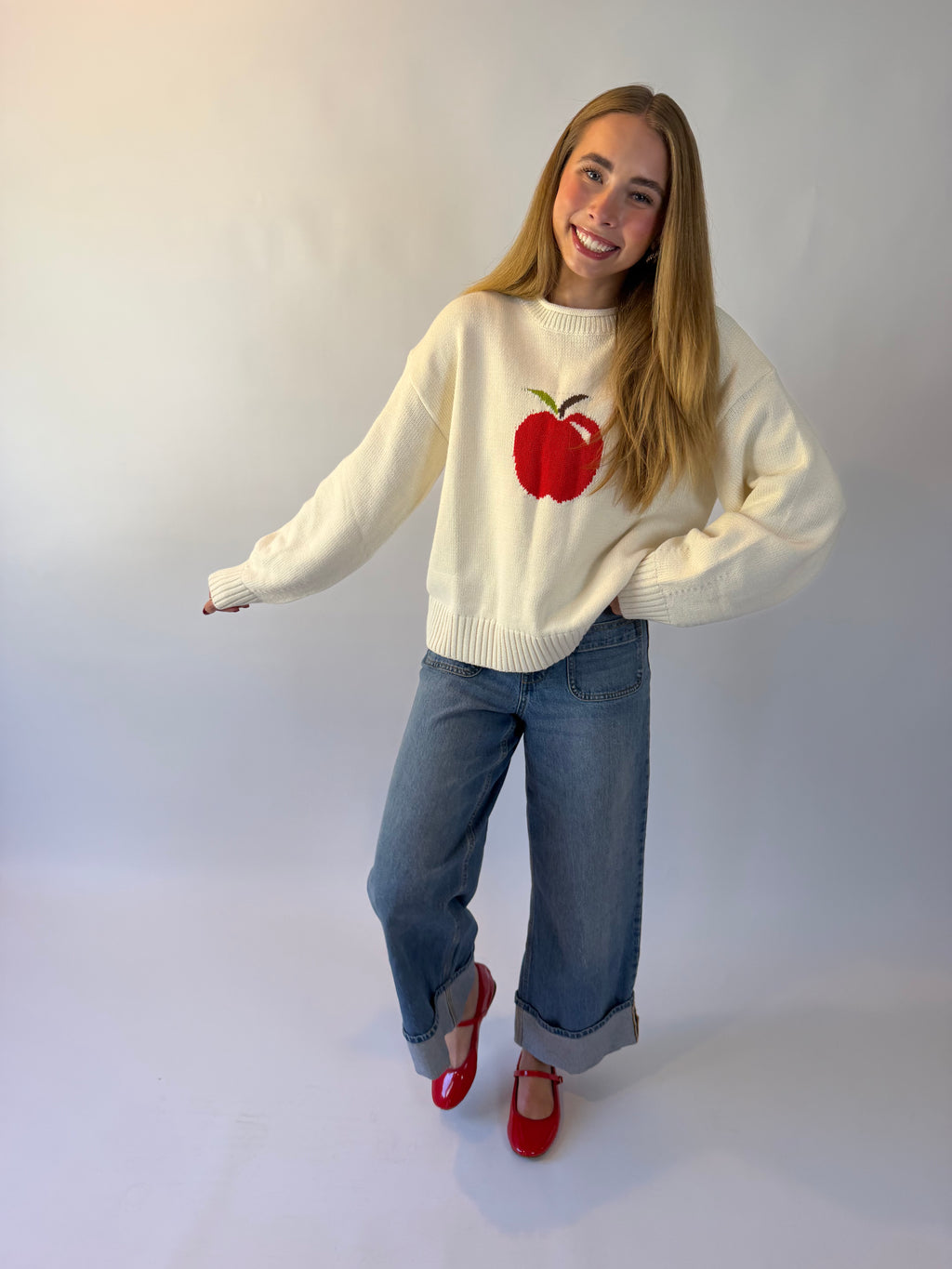 A young woman wearing a cream knit sweater with a red apple graphic on the chest, blue jeans, and red shoes, photographed against a plain light background.