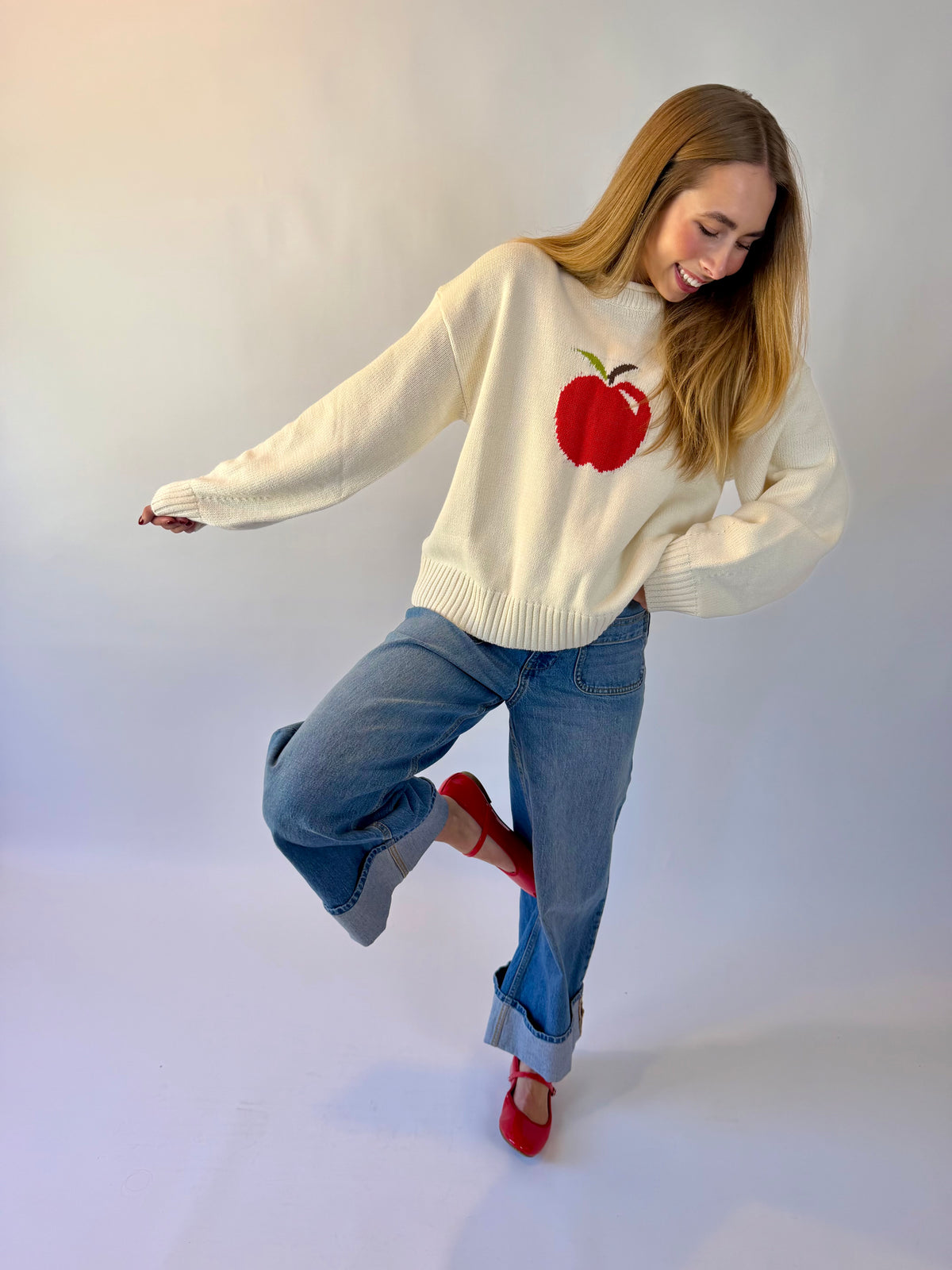 A woman posing in a cream-colored knit sweater with a red apple graphic on the chest, paired with blue jeans and red shoes, against a plain light gray studio background.