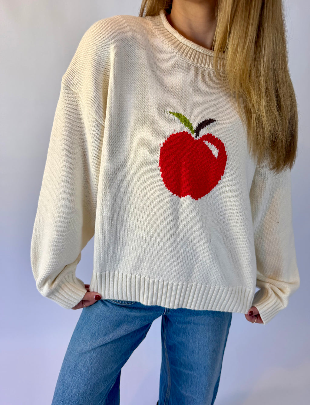 A person wearing an oversized cream knit sweater with a large red apple motif on the chest, paired with blue jeans, against a plain light background.