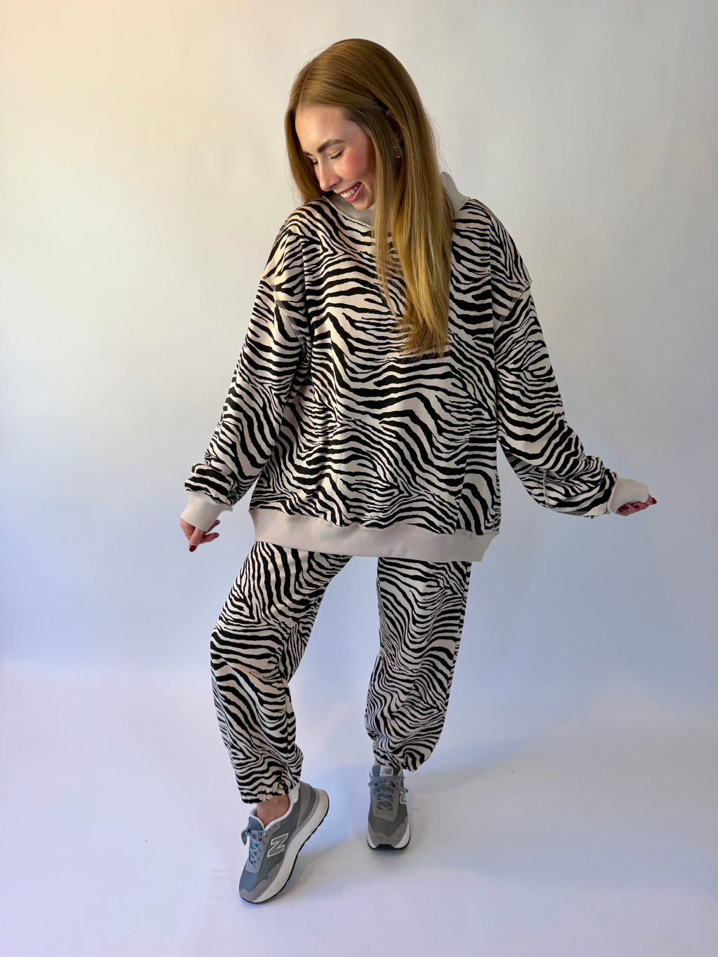 A woman modeling a matching zebra-print fleece loungewear set, featuring a long-sleeve hoodie and jogger pants, paired with gray sneakers against a plain white studio background.