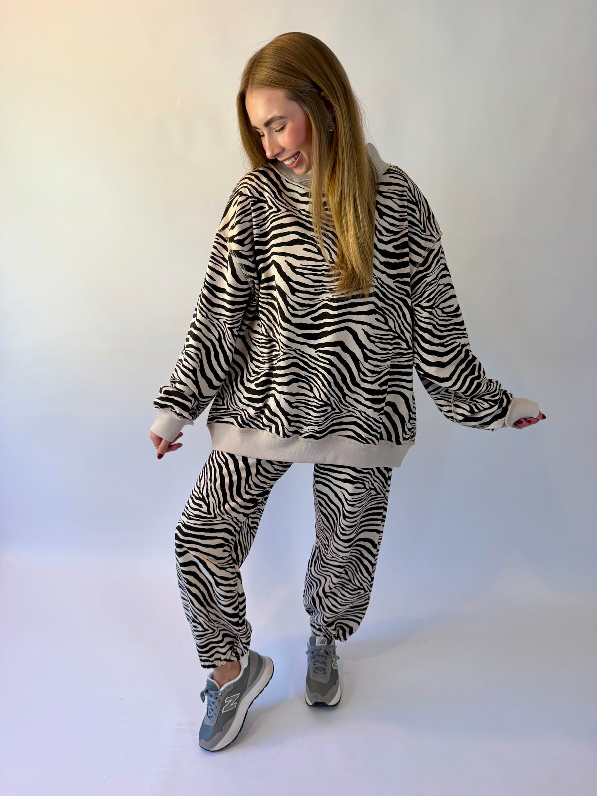 A woman modeling a matching zebra-print fleece loungewear set, featuring a long-sleeve hoodie and jogger pants, paired with gray sneakers against a plain white studio background.