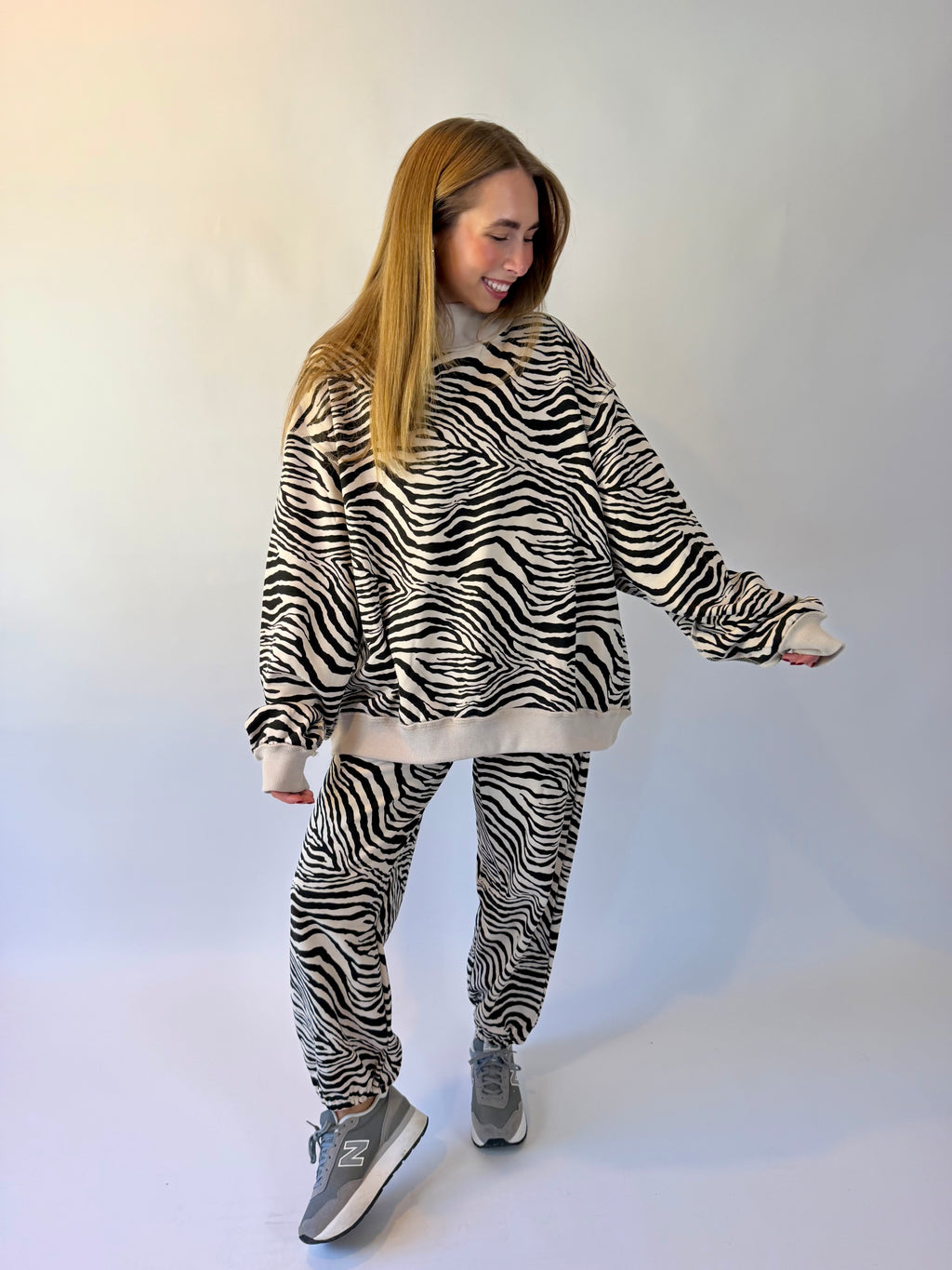 A woman modeling a matching zebra-print fleece loungewear set, featuring a cropped sweatshirt and jogger pants, styled with gray New Balance sneakers against a plain white studio background.