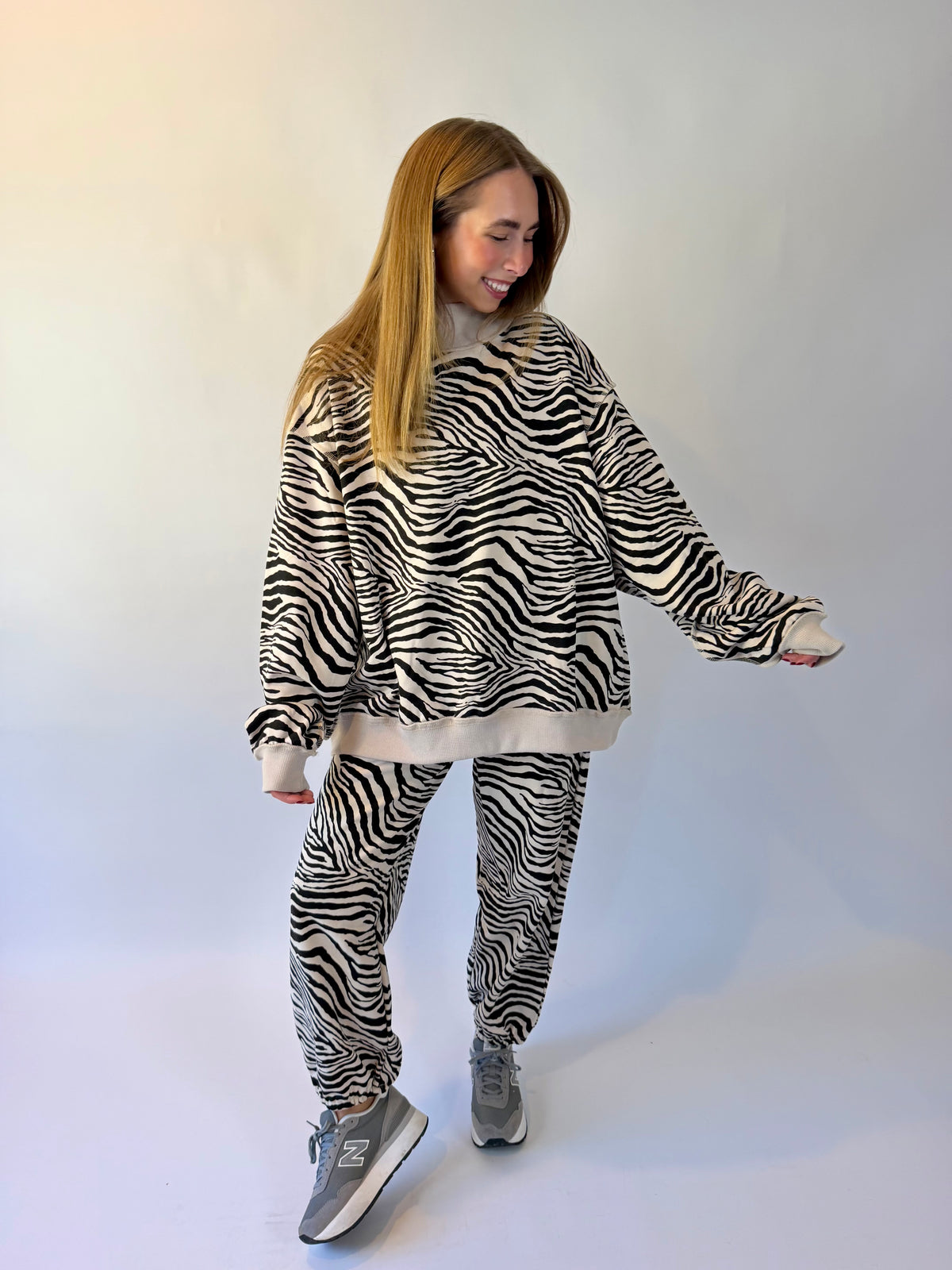 A woman modeling a matching zebra-print fleece loungewear set, featuring a cropped sweatshirt and jogger pants, styled with gray New Balance sneakers against a plain white studio background.