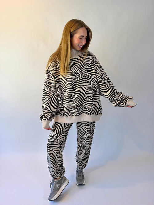 A woman modeling a matching zebra-print fleece loungewear set, featuring a cropped sweatshirt and jogger pants, styled with gray New Balance sneakers against a plain white studio background.
