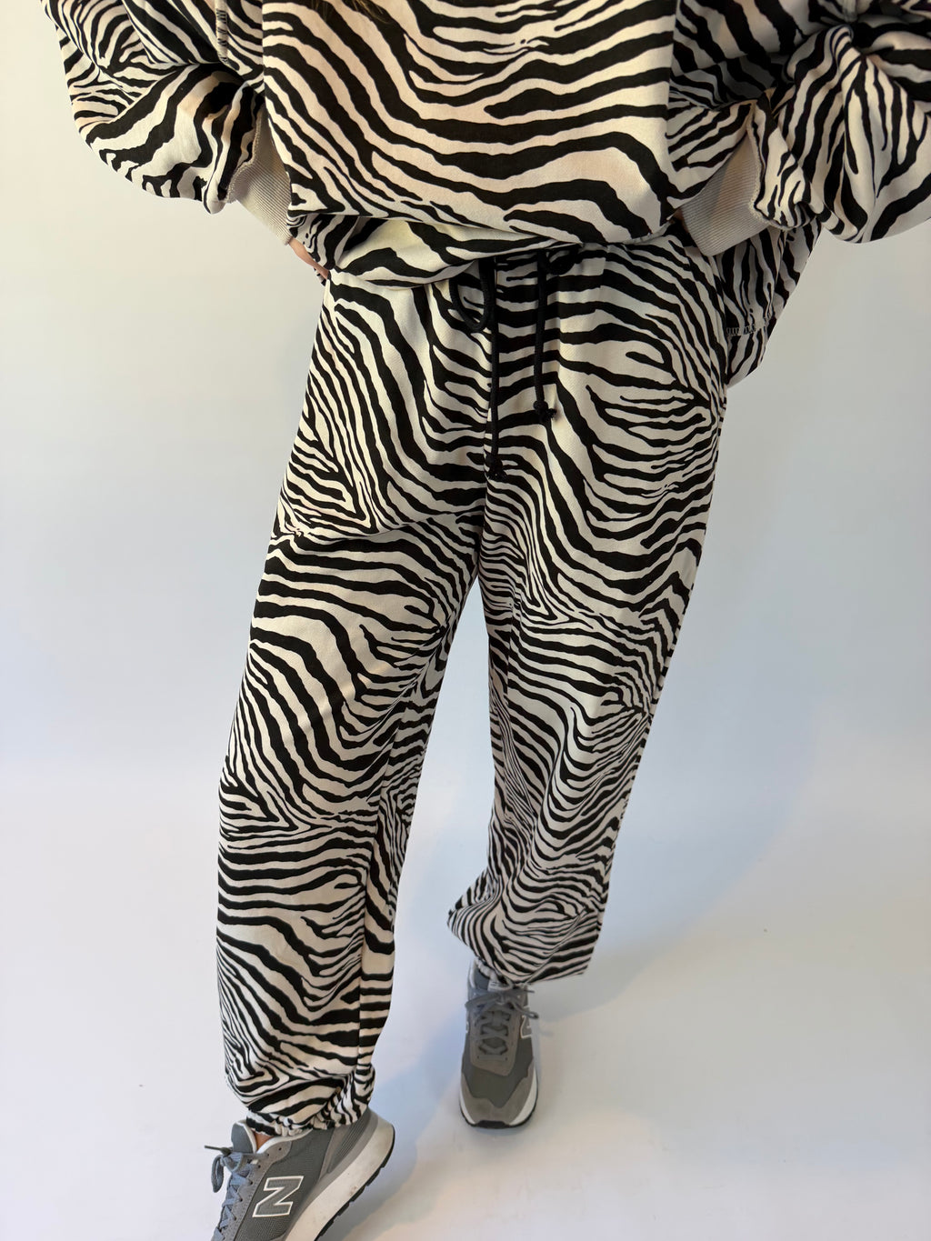 A person wearing zebra-print pants and a matching top, paired with gray New Balance sneakers, photographed against a plain white wall.