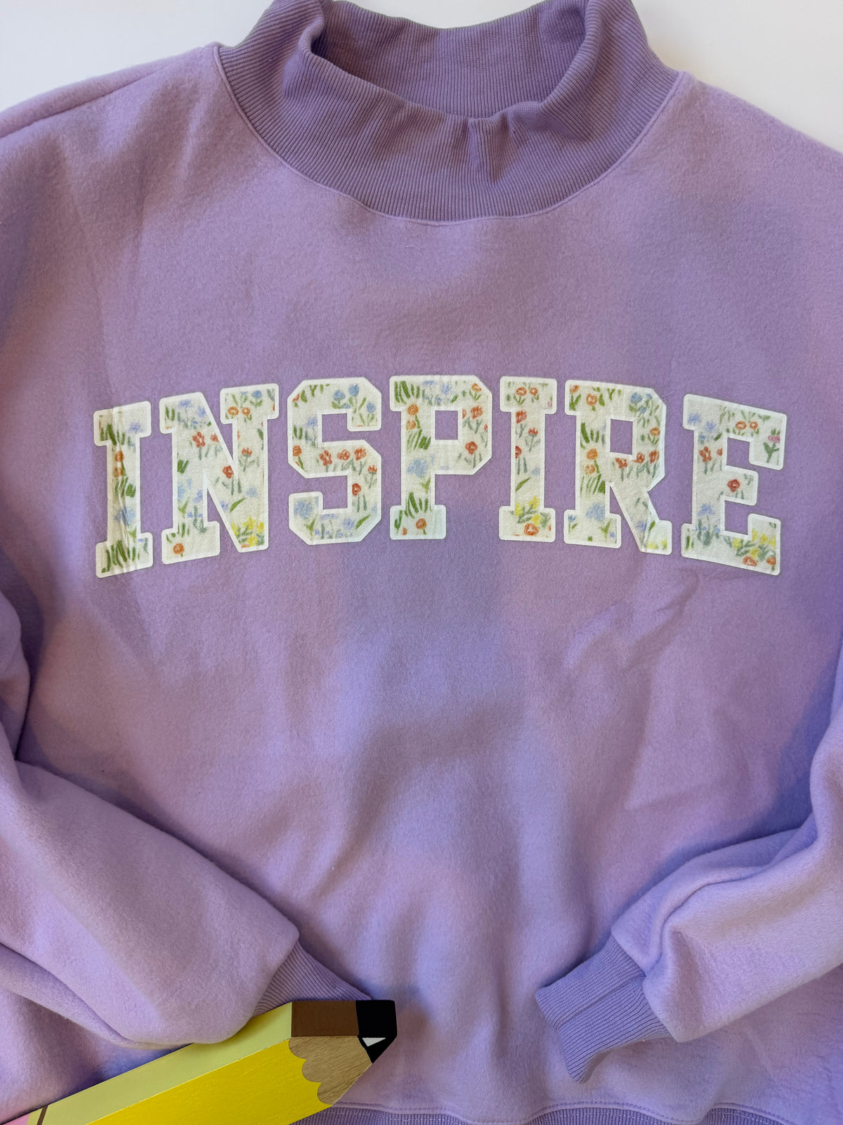 A lavender purple crewneck sweatshirt featuring the word 'INSPIRE' printed across the chest in arched varsity-style letters filled with a delicate floral and grass pattern. The sweatshirt is laid flat on a white surface with a yellow pencil partially visible at the bottom.