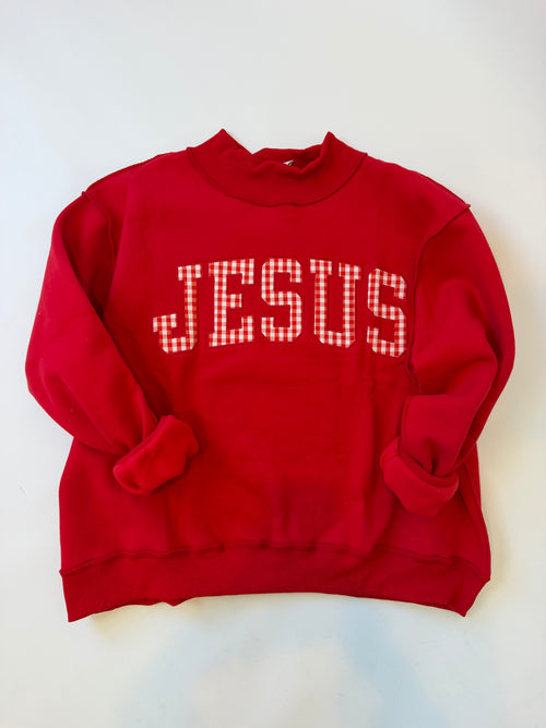 A red crewneck sweatshirt with the word 'JESUS' printed vertically on the front in a white and red gingham checkered pattern, laid flat on a white background.