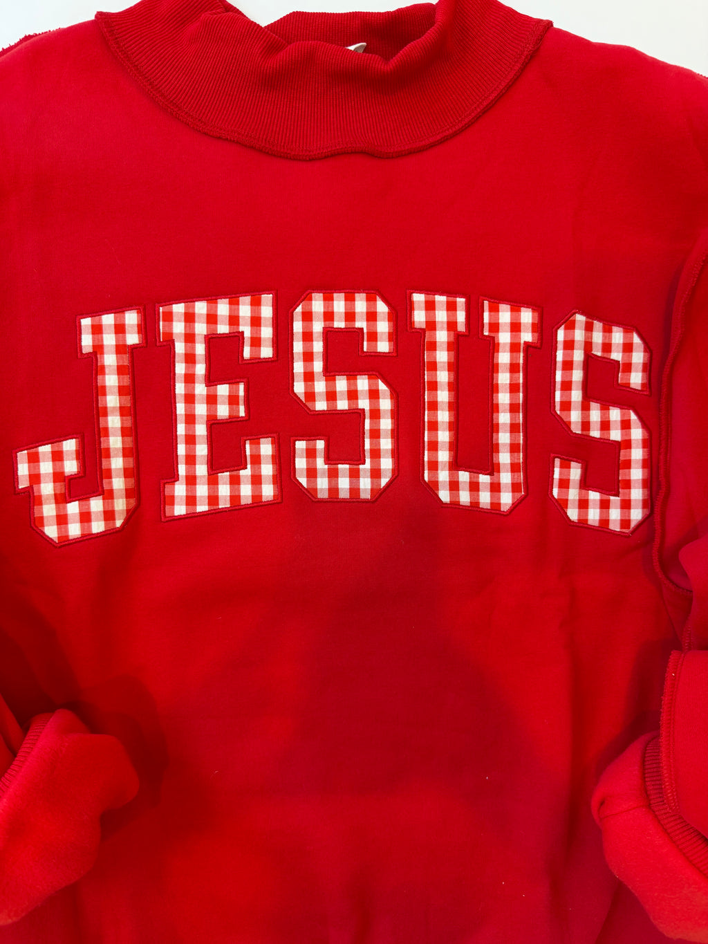 A red crewneck sweatshirt featuring the word 'JESUS' in large varsity-style block letters across the chest. The letters are filled with a red and white gingham checkered pattern, which contrasts with the solid red body of the garment.