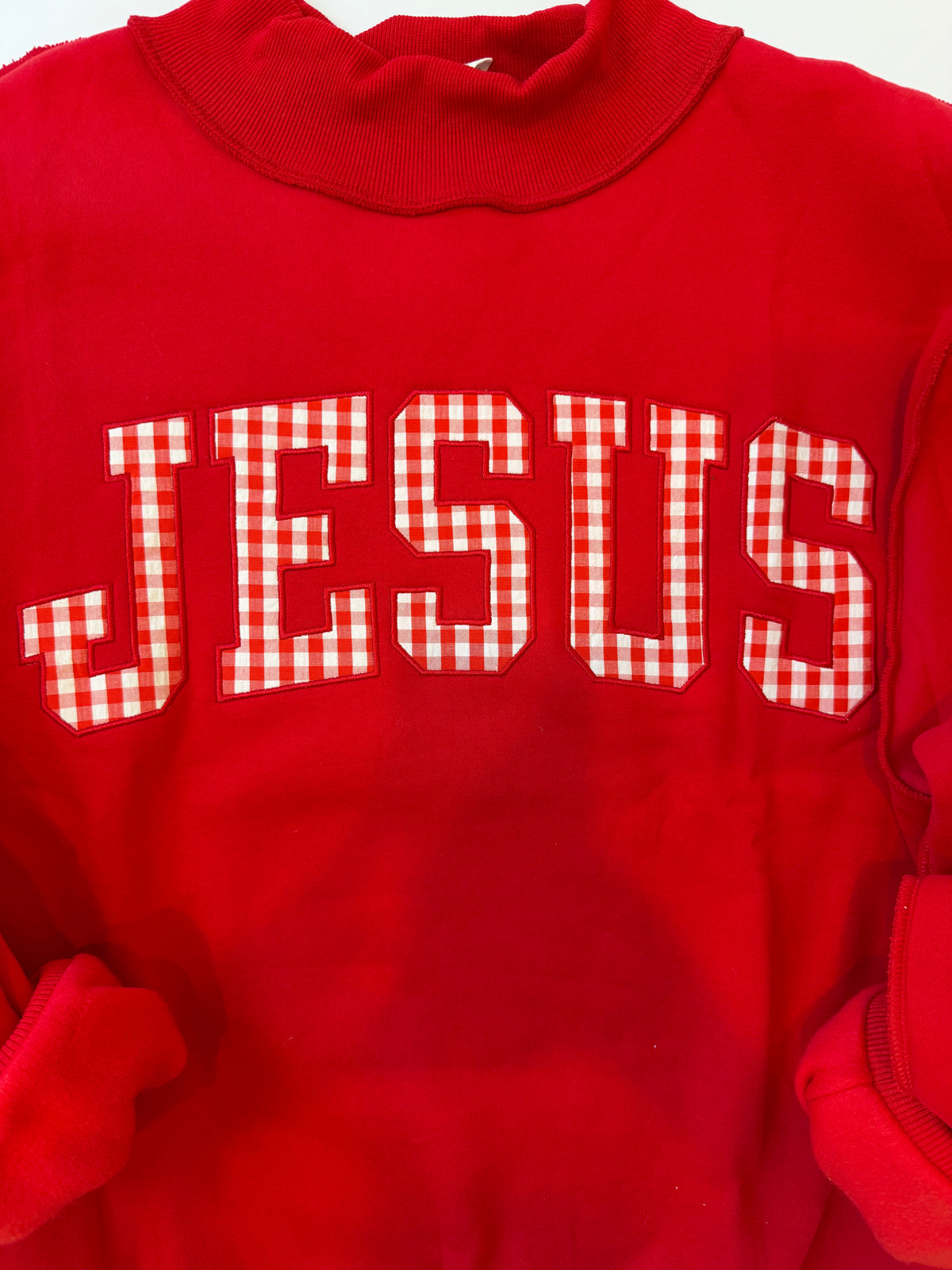 A red crewneck sweatshirt featuring the word 'JESUS' in large varsity-style block letters across the chest. The letters are filled with a red and white gingham checkered pattern, which contrasts with the solid red body of the garment.