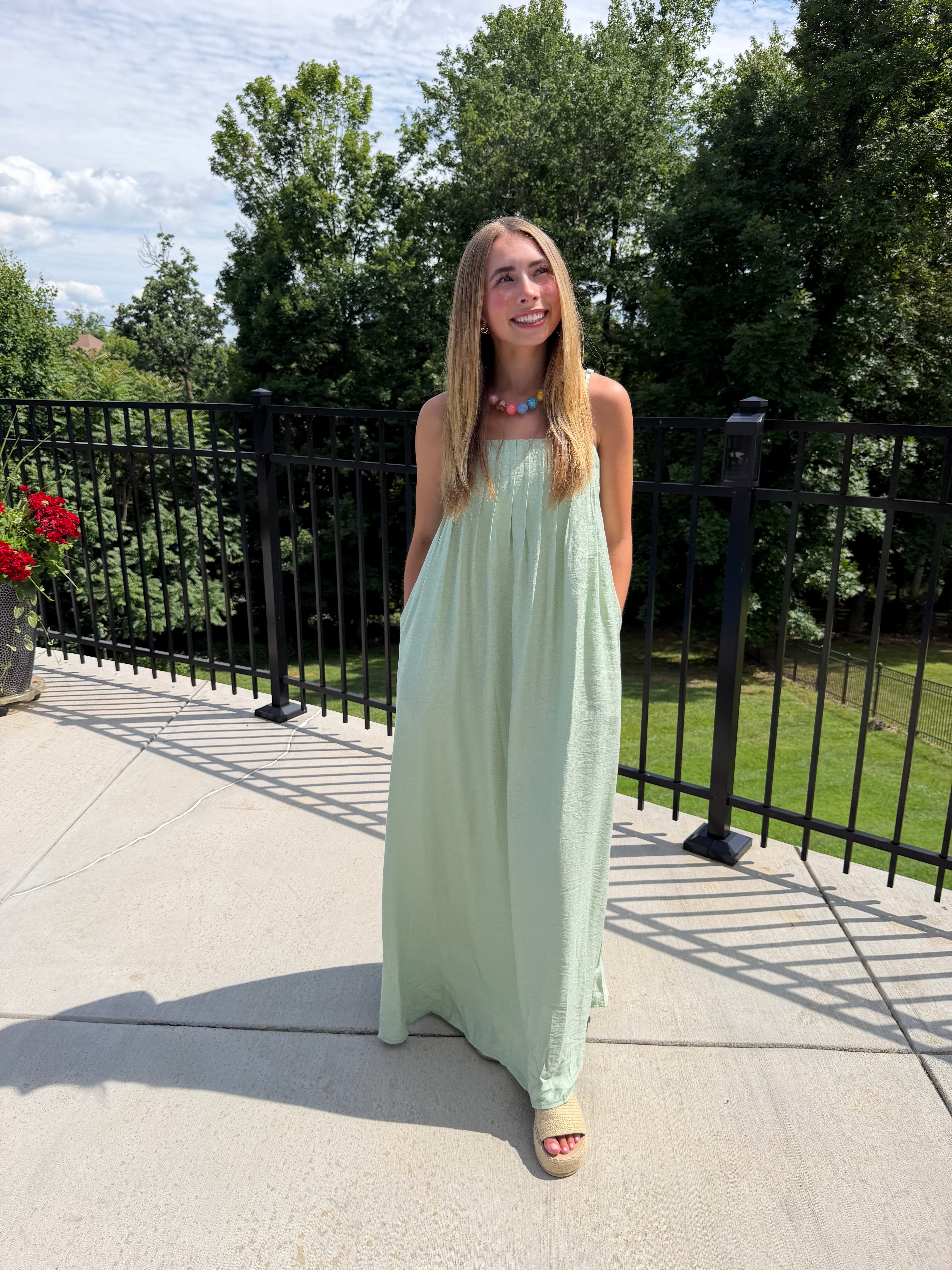 Woman in a light green dress standing outdoors with trees and a fence in the background