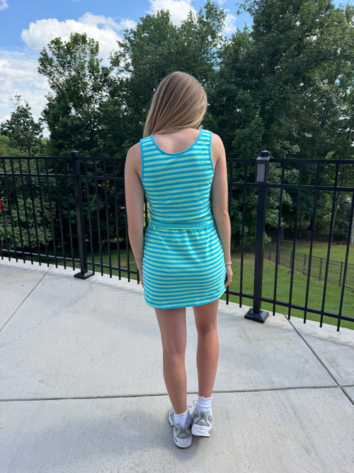 Person wearing a blue and green striped dress standing on a concrete surface with trees in the background.