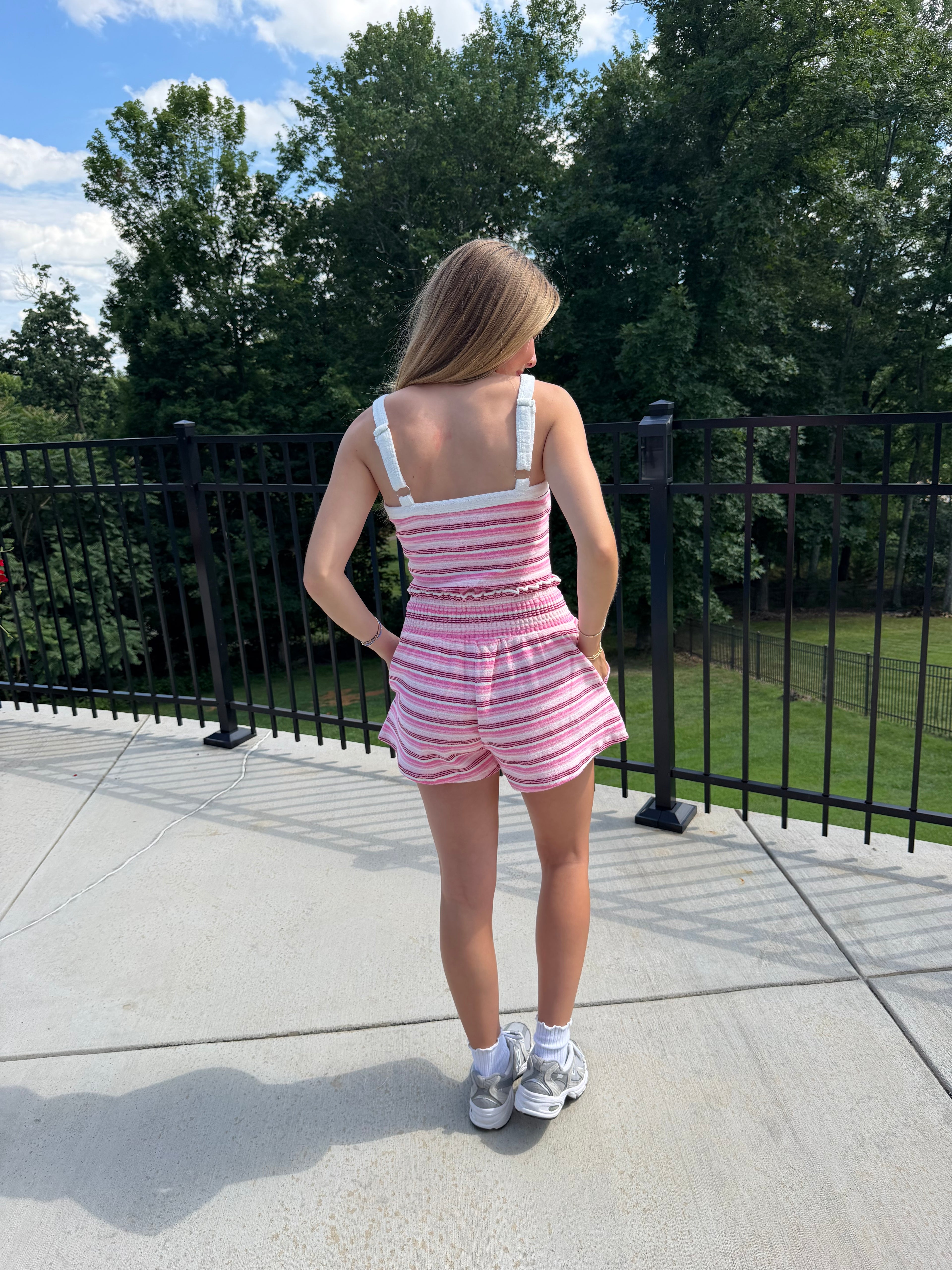 Person in a pink and white striped dress standing on a sidewalk with trees and a fence in the background.
