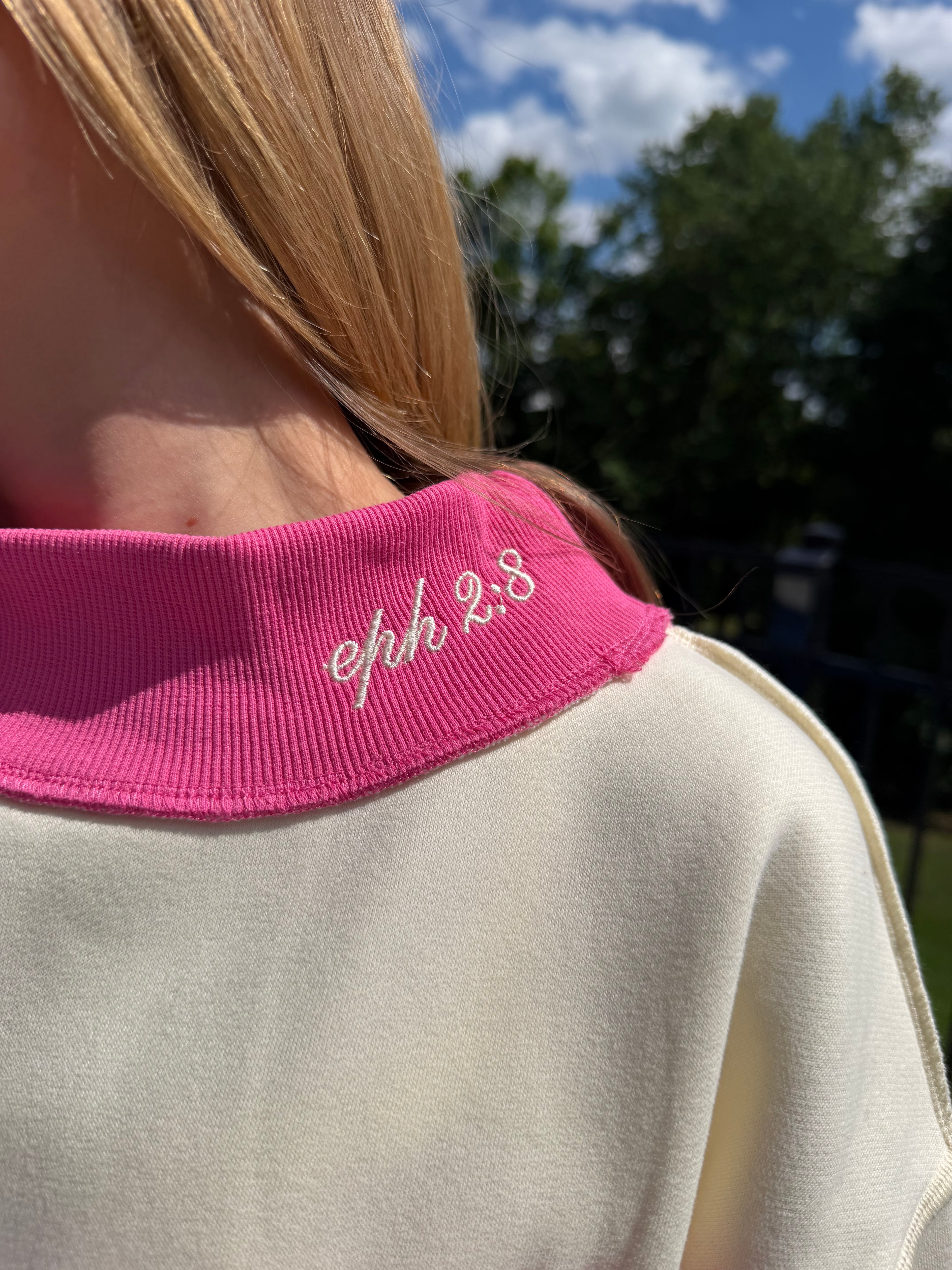 Close-up of a pink collar with 'eph 2:8' embroidery on a beige garment against a blurred outdoor background.