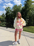Person wearing a cream-colored sweatshirt with text, pink shorts, and white sneakers on a sunny day outdoors.